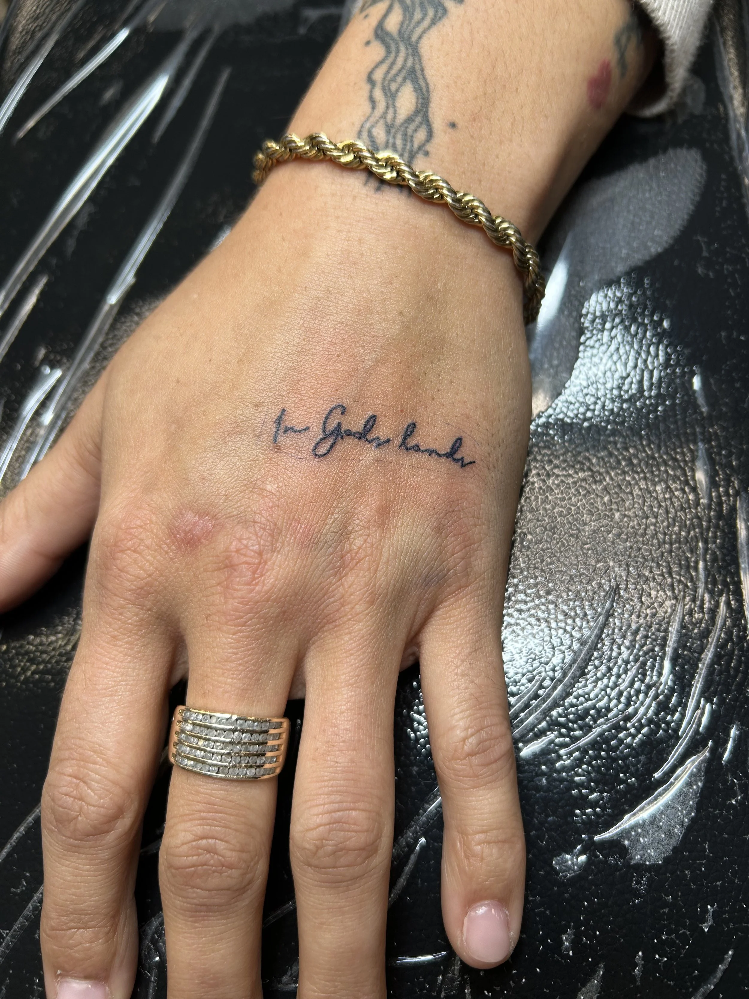 A hand with a black ink tattoo that says 'to Golden hander,' a gold twisted bracelet, and a silver ring with multiple rows of small diamonds. The background is black with a textured surface.