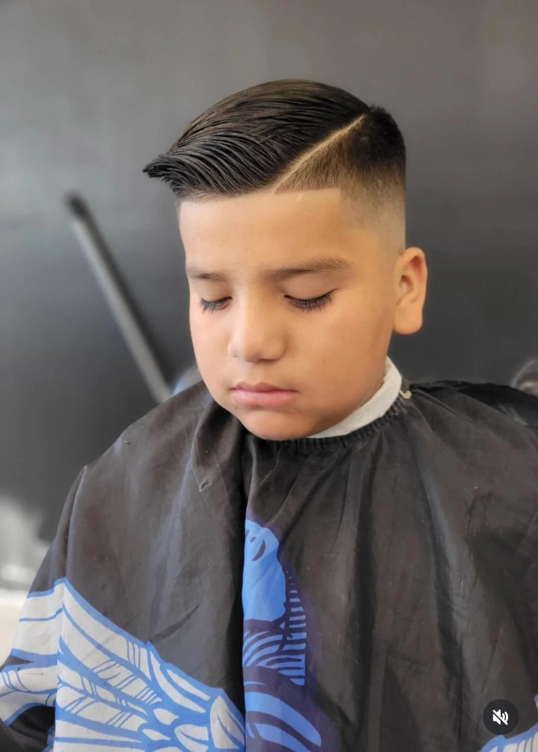 Young boy with a sleek, side-part hairstyle sitting in a barber's chair, wearing a black cape with a blue and white design, with eyes closed.