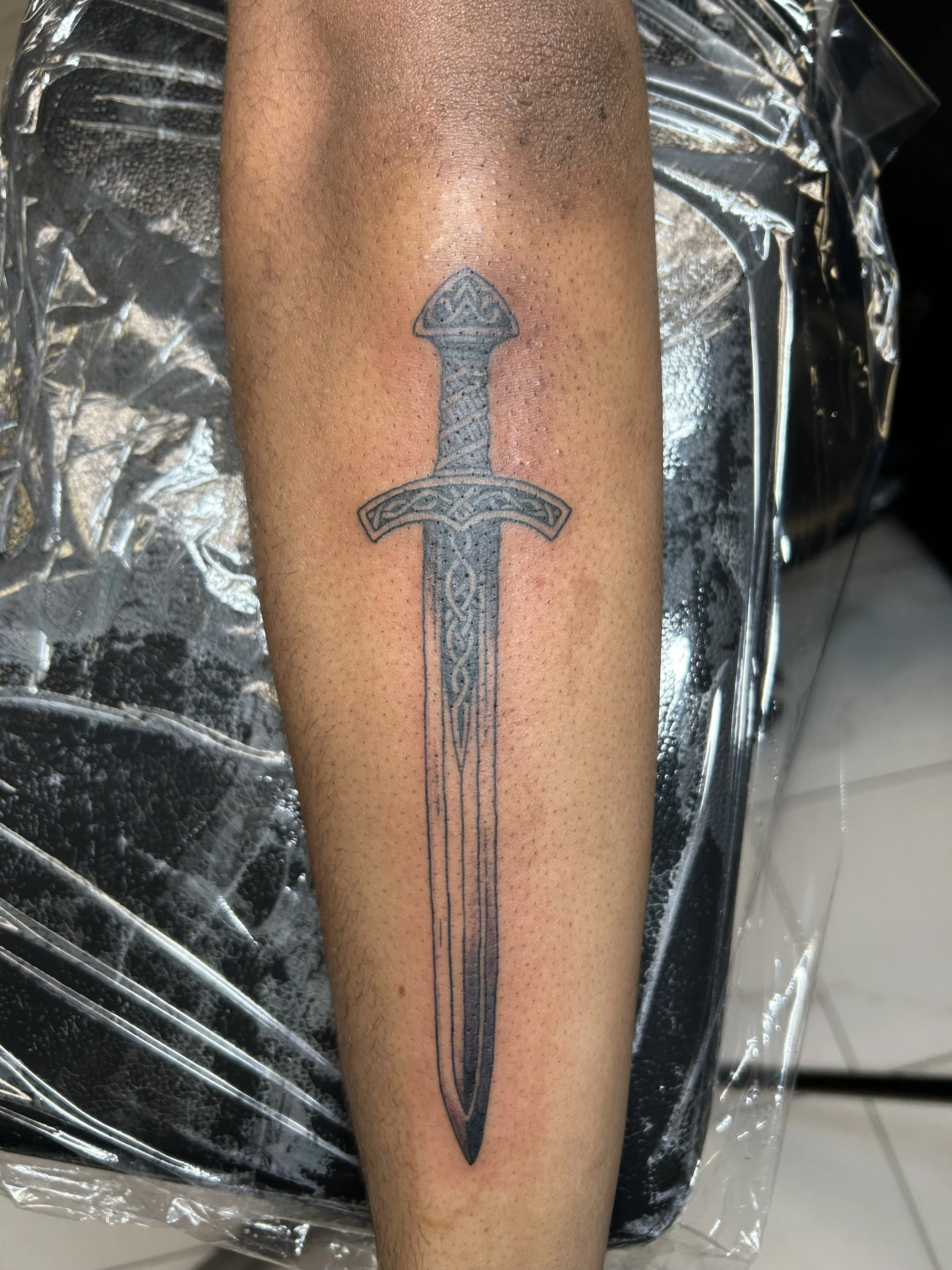 Black outline tattoo of a sword with intricate Celtic knot designs on a person's forearm.