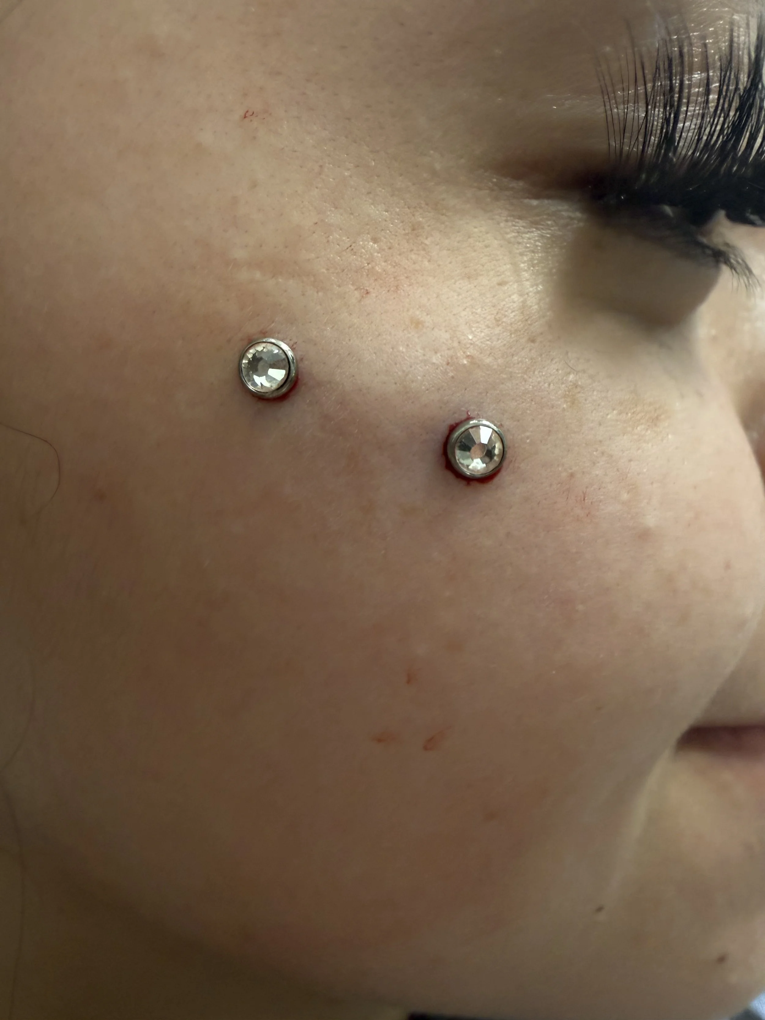 Close-up of a person's cheek with two facial piercings featuring clear gemstones, and part of the eye with long eyelashes visible.