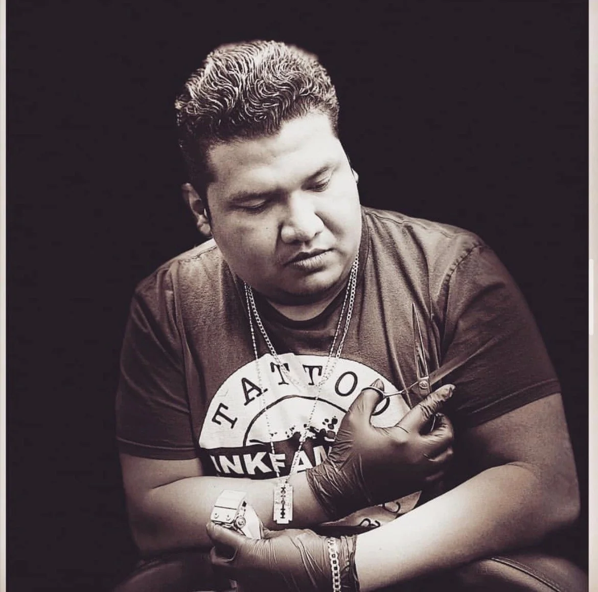 Black and white photo of a man with short curly hair, wearing a t-shirt with a graphic that reads 'Tattoo Ink' and multiple chains around his neck. He is wearing a tattoo needle on his left arm and black gloves, holding a tattoo needle in his right hand. The background is dark.