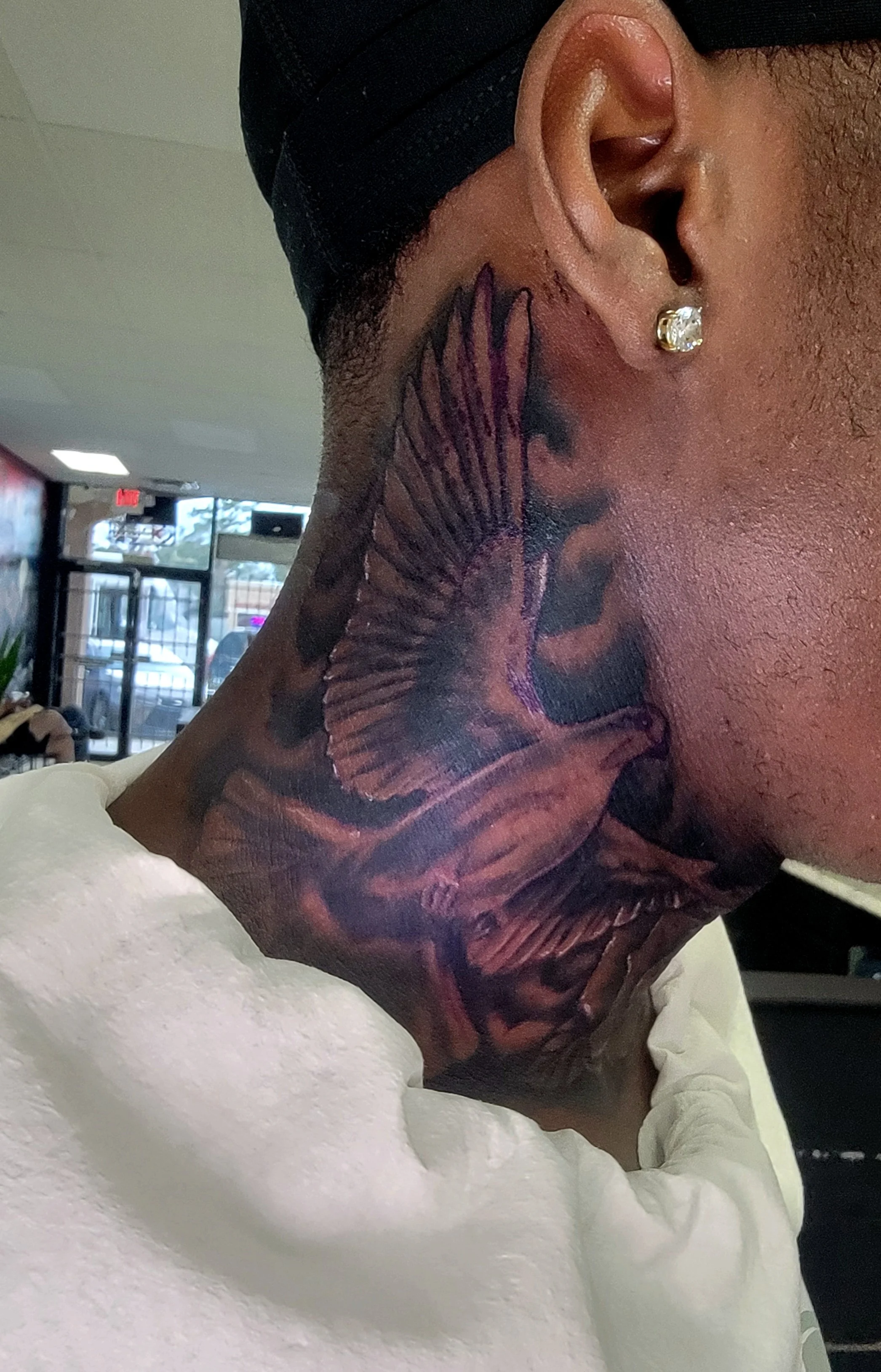 Close-up of a person's neck and ear showing a large tattoo and wearing a diamond earring, with a blurred background of a store interior.