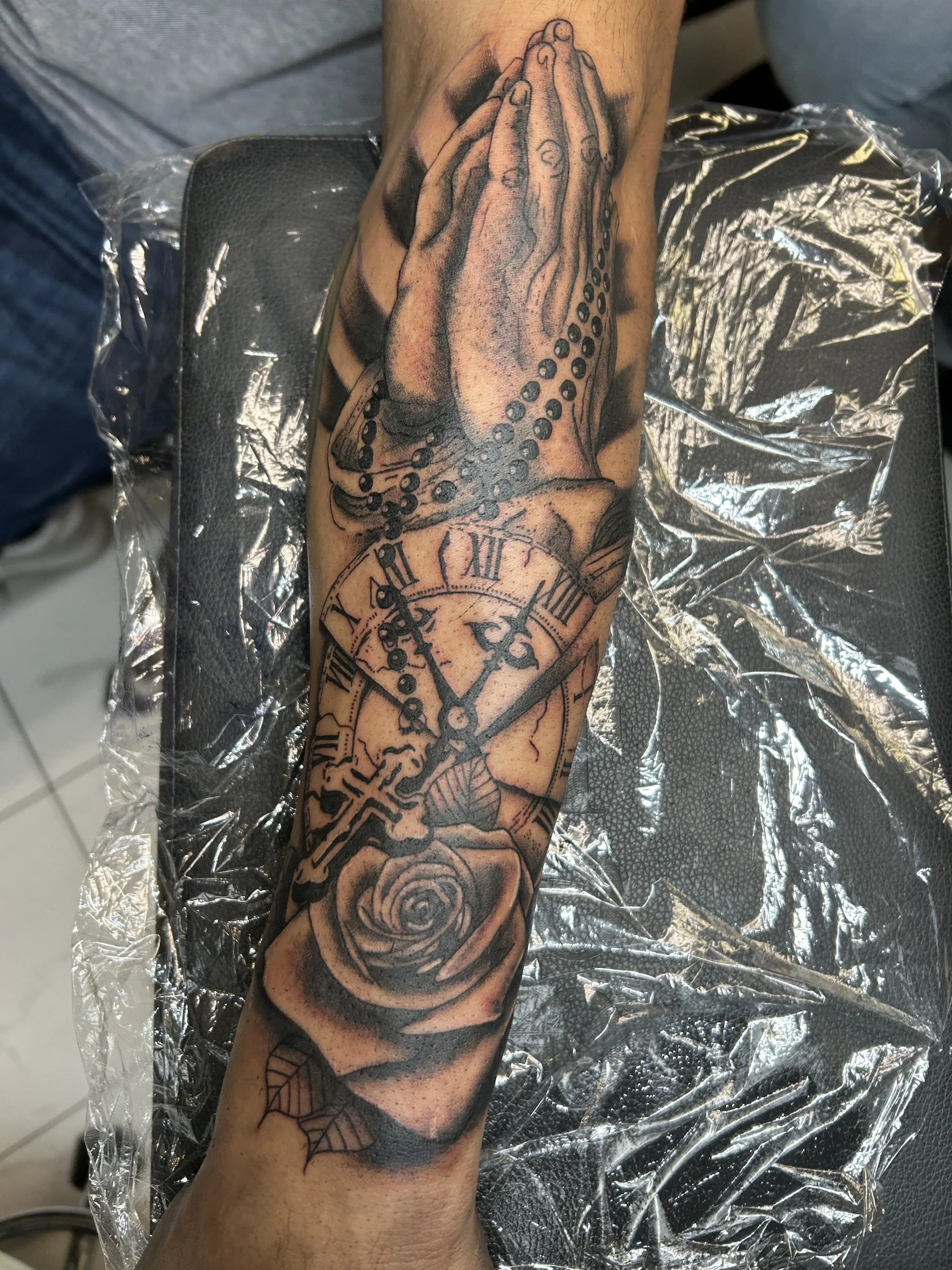 Tattoo of praying hands holding rosary beads, with a clock face showing the time 10:07, and a rose at the bottom on a person's forearm.