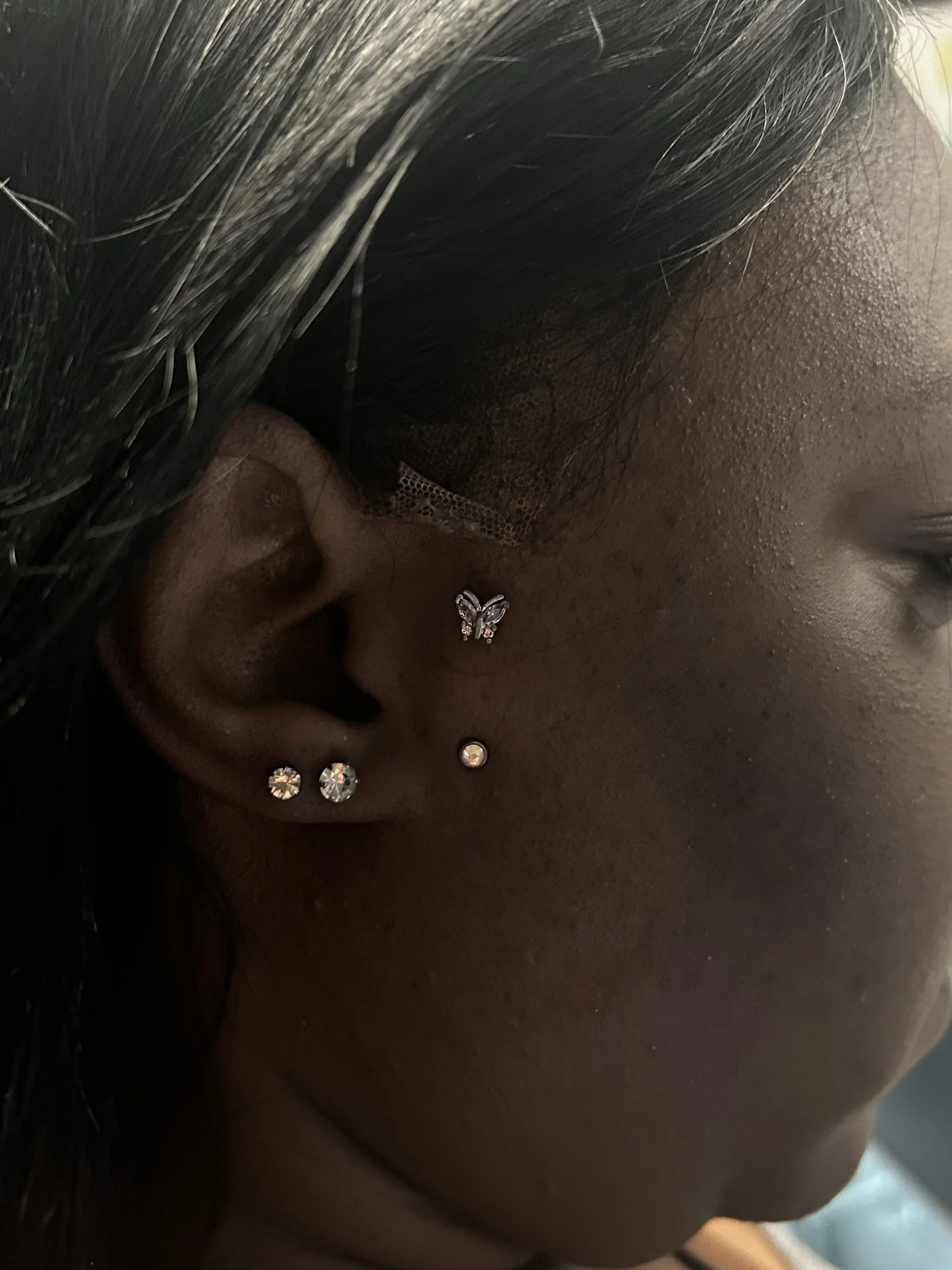 Close-up of a person's ear with three small diamond studs, a butterfly-shaped earring, and a black lace face mask.