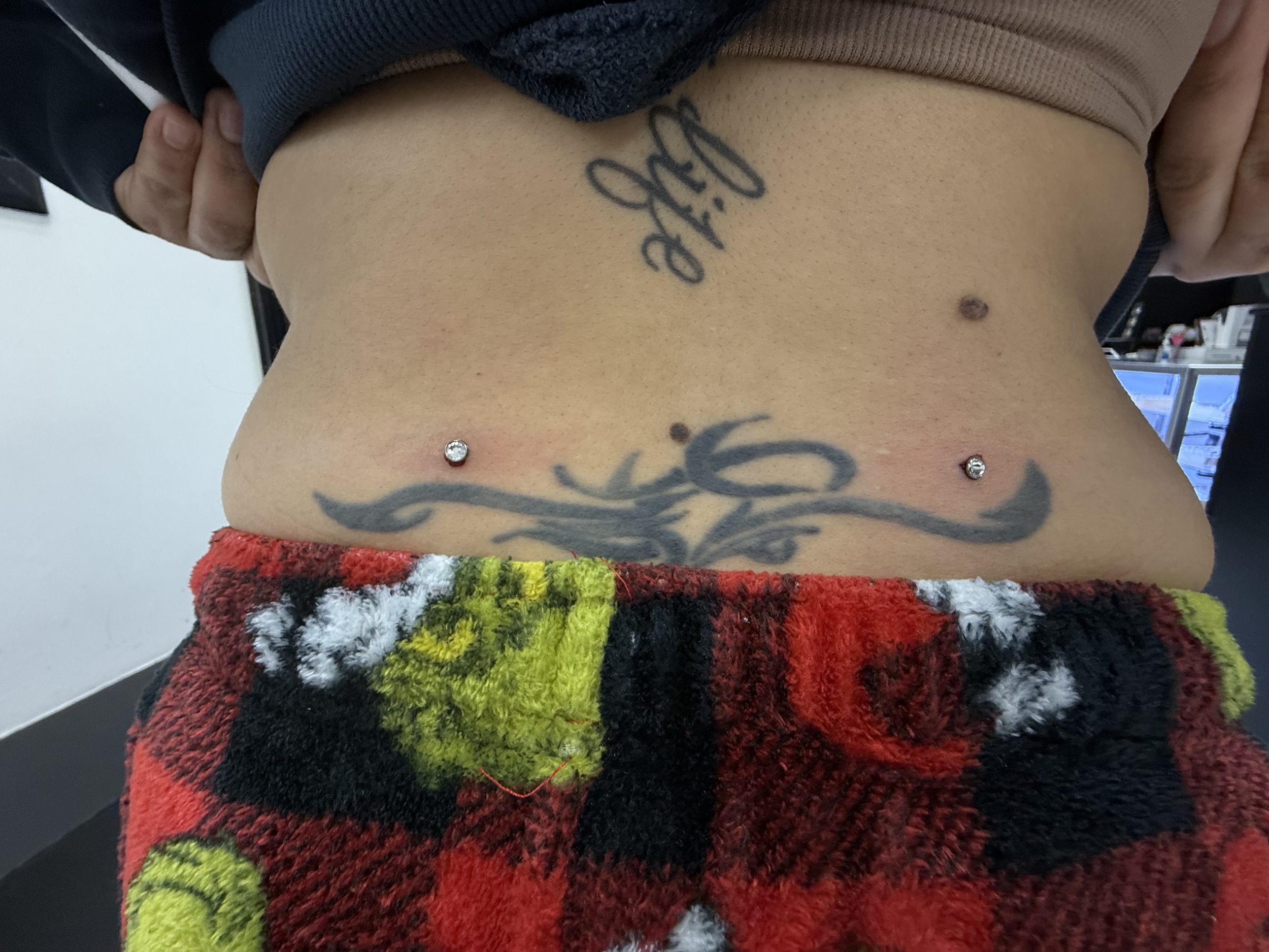 A person is lifting their shirt to reveal a tattoo on their abdomen, with a colorful, fluffy blanket below and piercings on their belly button.