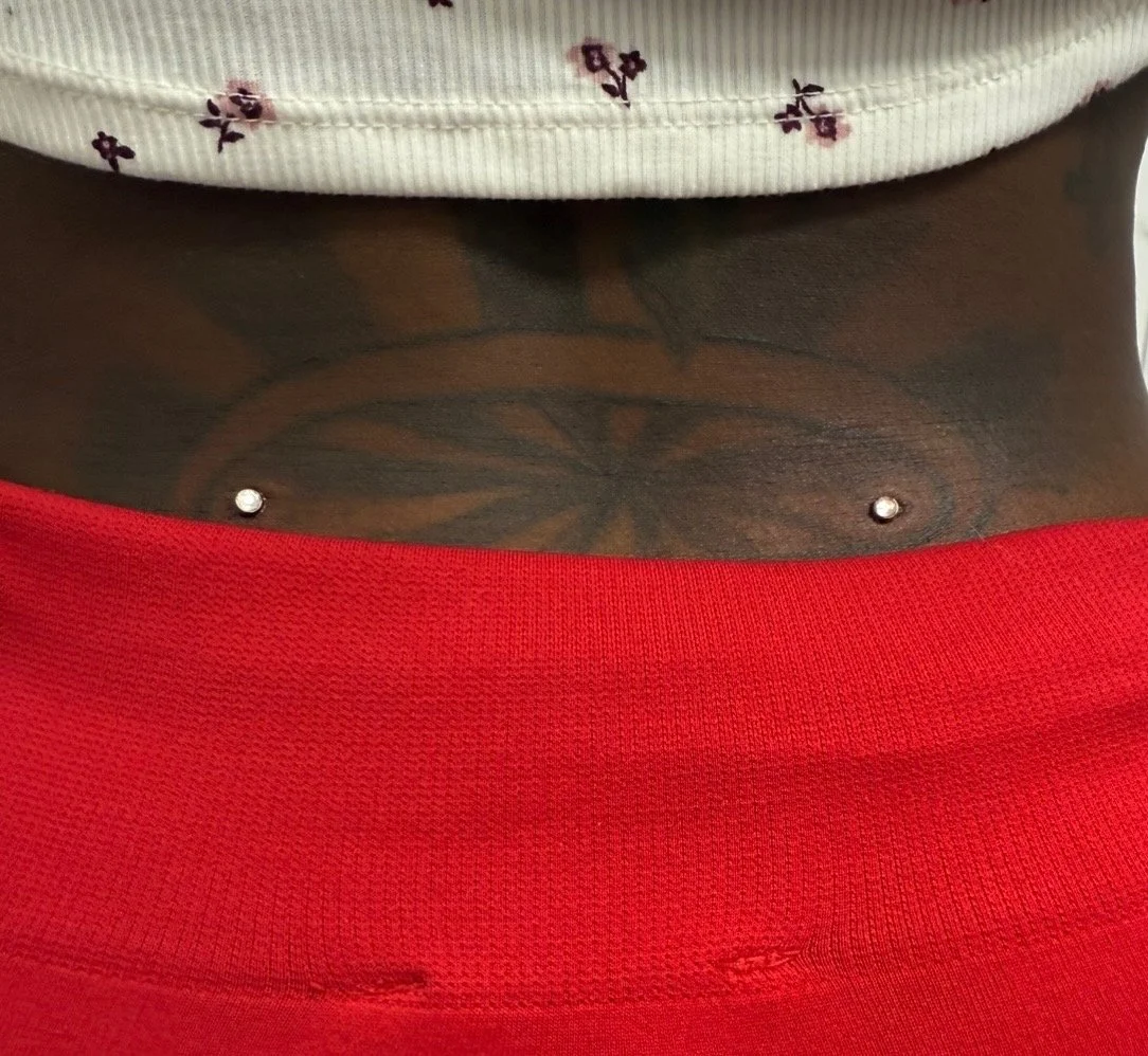A person's lower abdomen showing a tattoo and two belly button piercings, wearing red pants and a white shirt with small floral patterns.