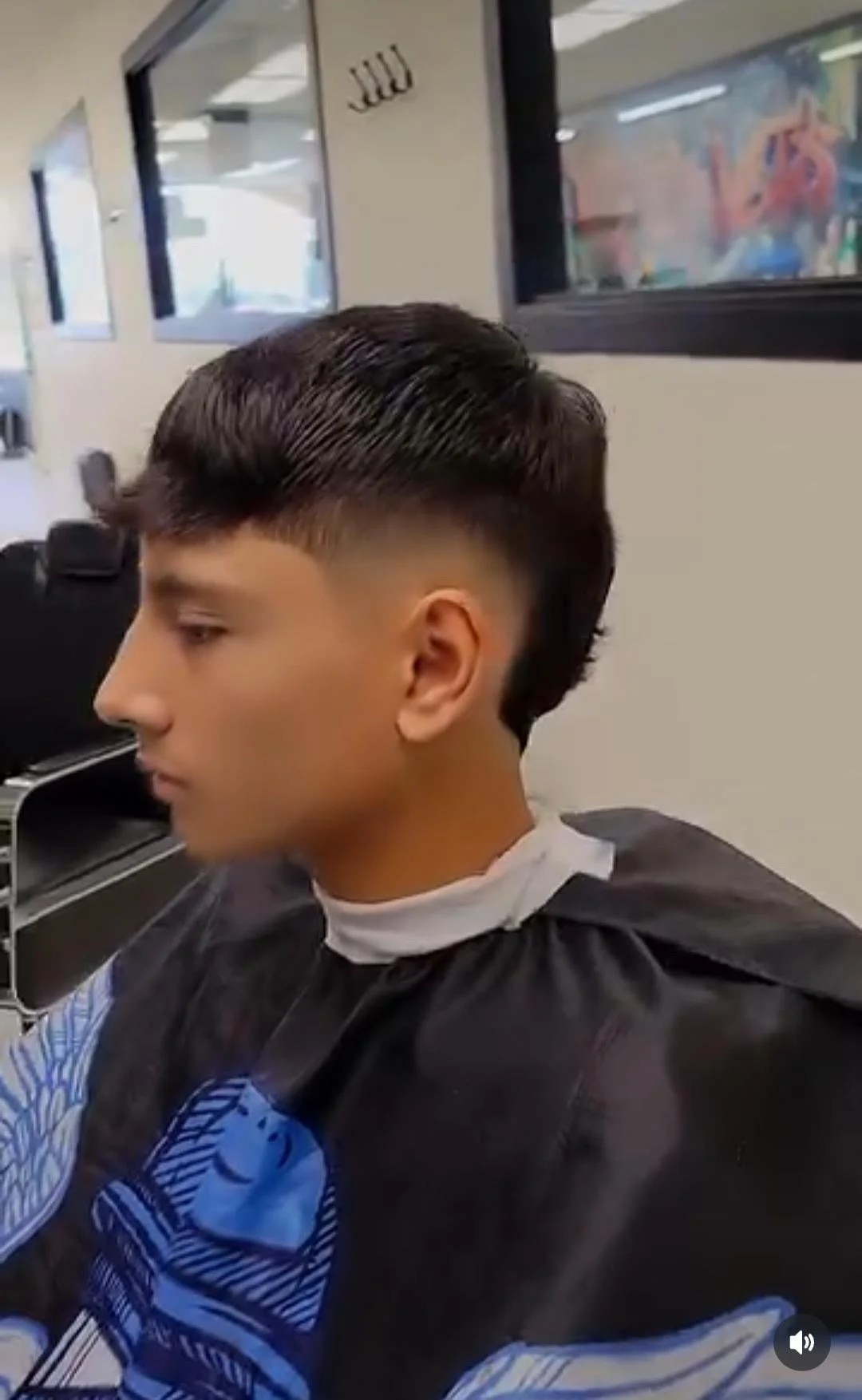 Young man with a modern hairstyle, shaved sides and combed back top, sitting in a barbershop wearing a black barber cape with blue and white patterns, getting a haircut.
