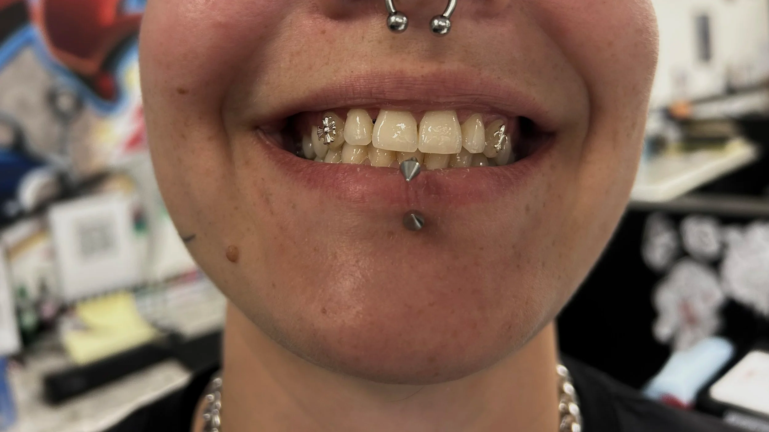 Close-up of a person’s face showing a broad smile with tattoos, piercings, and decorated teeth, including a flower and star designs on the teeth and metallic piercings on the lips and nose.