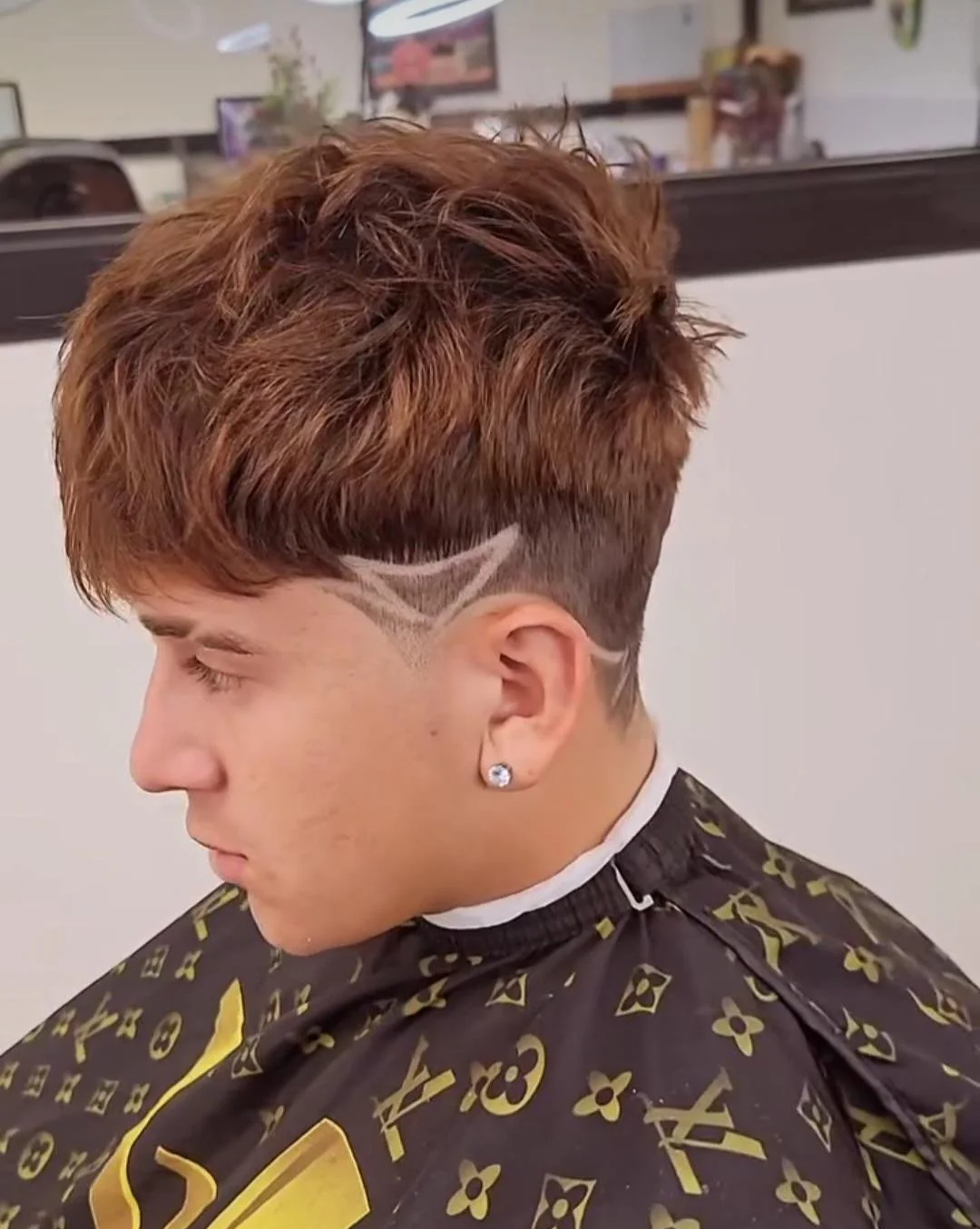 Young man with a textured, reddish-brown hairstyle featuring a geometric shave design on the side of his head, wearing a black and gold patterned cape at a hair salon.