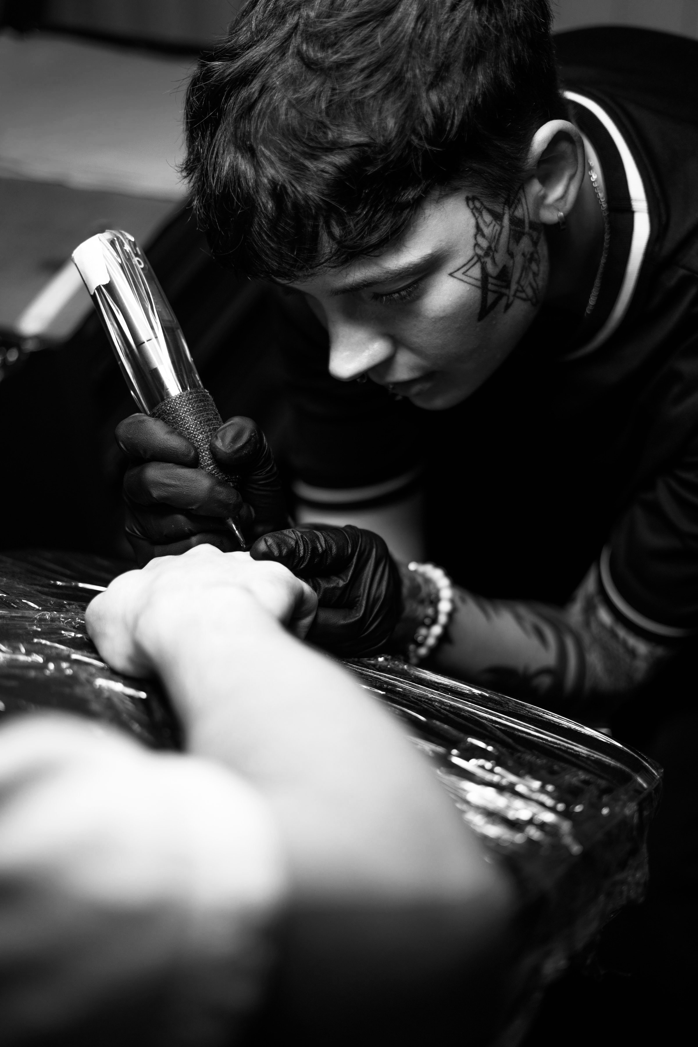 A person with short hair and tattoos on their neck, wearing black gloves, intensely drawing or tattooing on a surface with a tattoo machine or pen. The scene is in black and white.