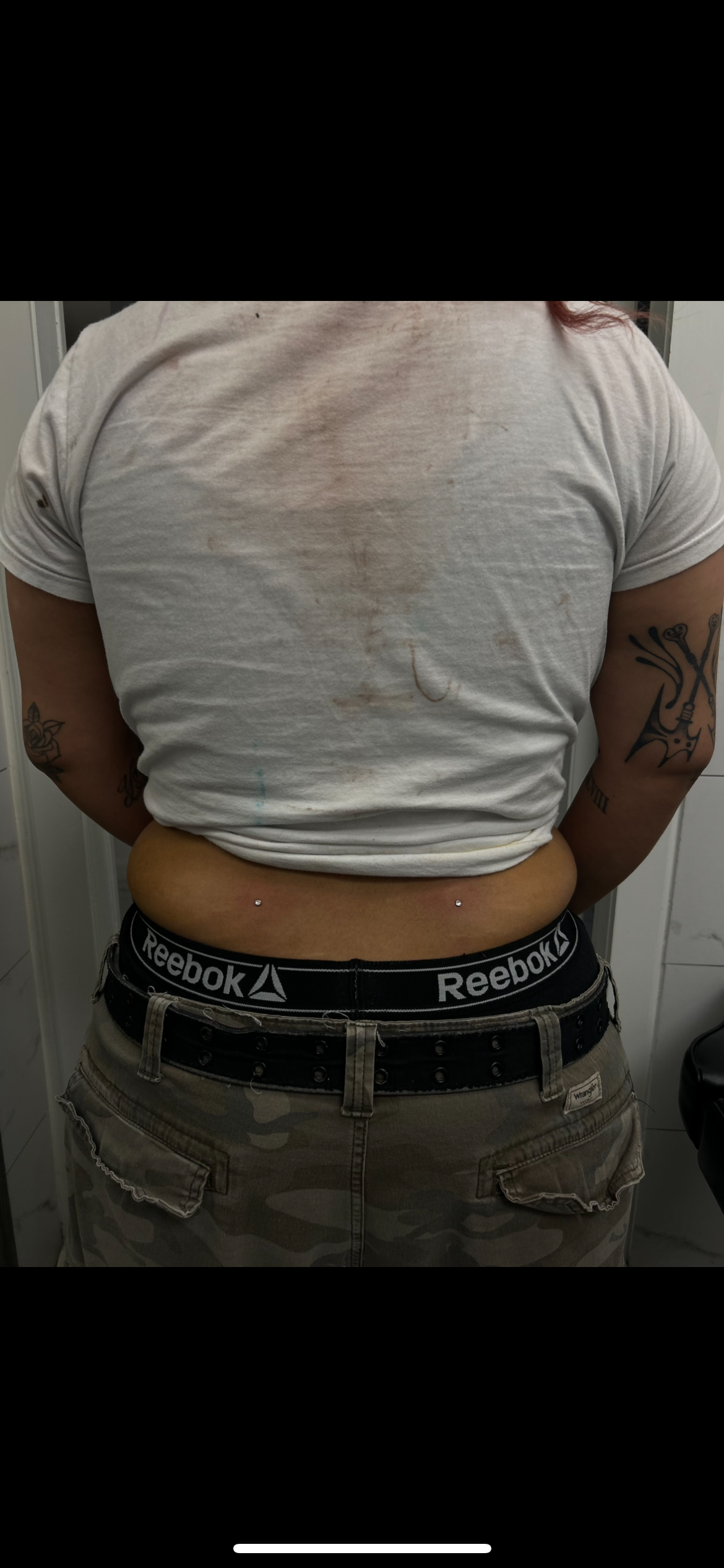 A person standing with a white t-shirt and camouflage shorts, showing a flat stomach with two piercing on the lower abdomen, tattoos on both arms, and a dresser in the background.