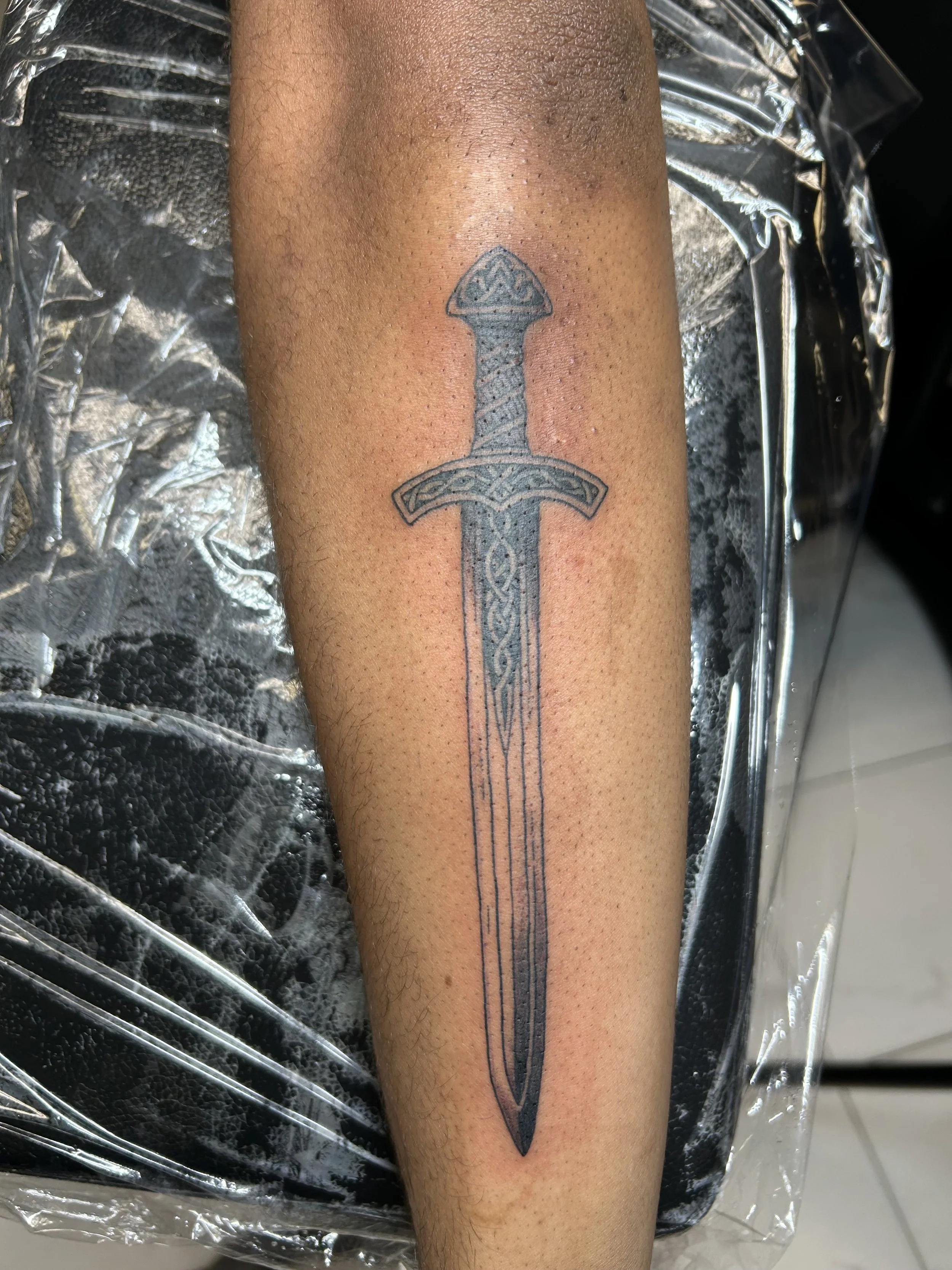 Freshly tattooed sword design on person's forearm with intricate details and a Celtic knot pattern in the hilt, surrounded by foam and plastic wrapping in a tattoo studio.