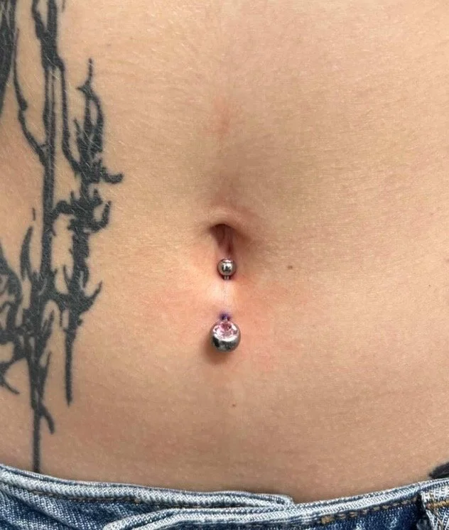 A close-up of a person's abdomen showing a tattoo on the left side, and a belly button piercing with a pink gemstone on a silver barbell.