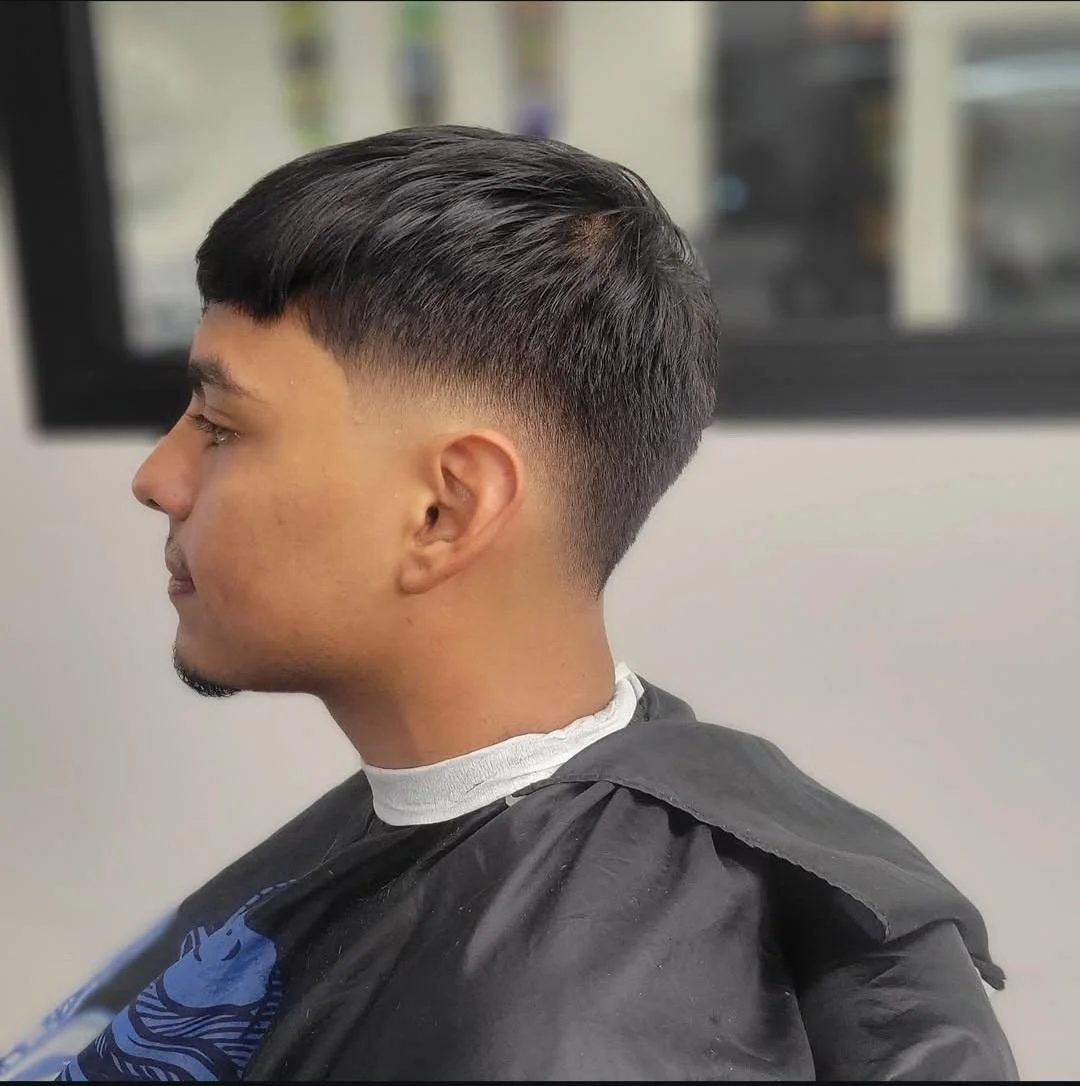 Side view of a young man with a fresh, faded haircut, wearing a black barber cape, sitting in a barber shop.