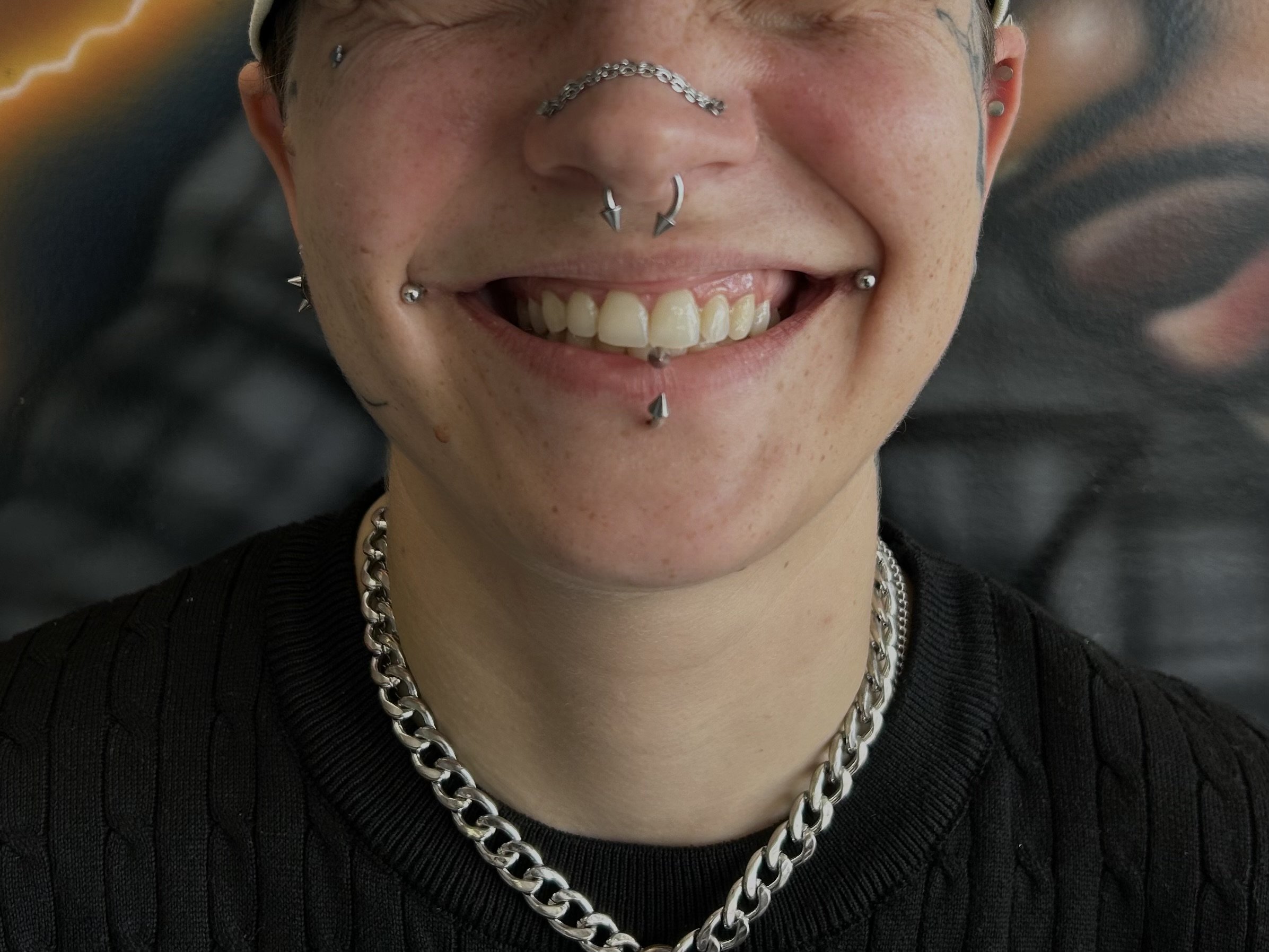 Close-up of a person's face with numerous facial piercings, including a chain across the nose, multiple ring piercings on lips, and studs on ears, wearing a chunky silver chain necklace and a black top.