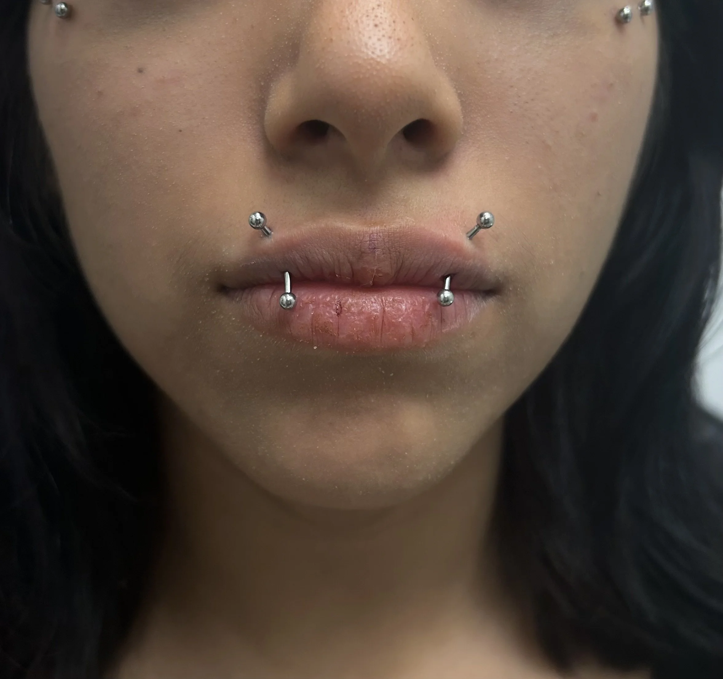 Close-up of a person's face showing nose, lips, and numerous facial piercings, including two on the nose and multiple on the lips and chin.
