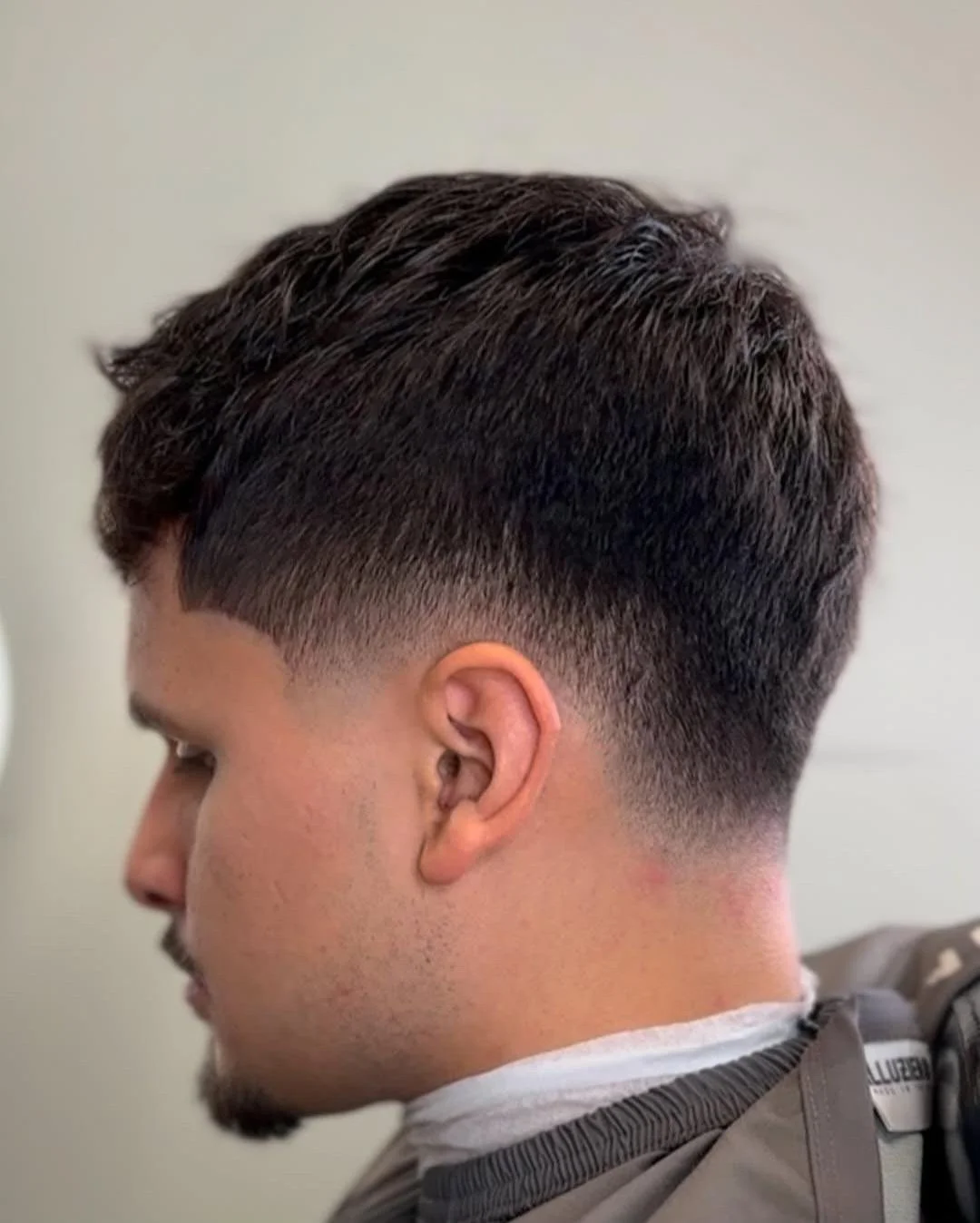 Side profile of a young man with a modern fade haircut, dark hair, and a light beard, wearing a barber cape.