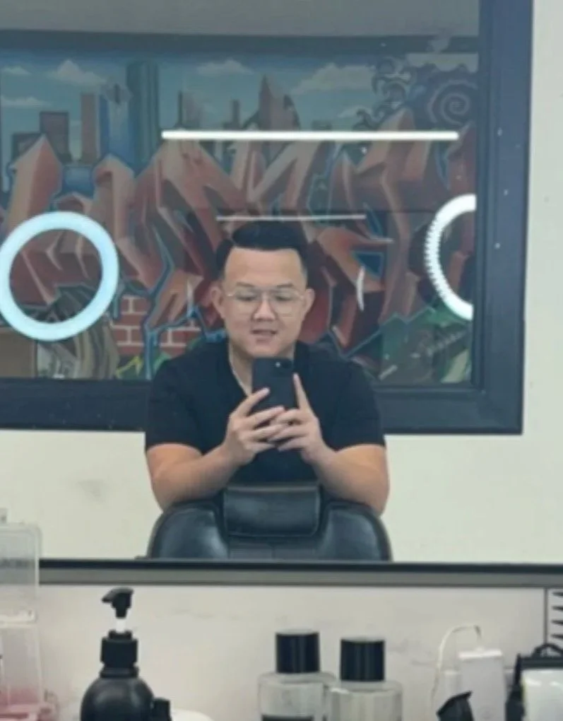 A young man takes a mirror selfie in a salon or barber shop, sitting on a chair in front of a mirror with colorful graffiti art in the background.