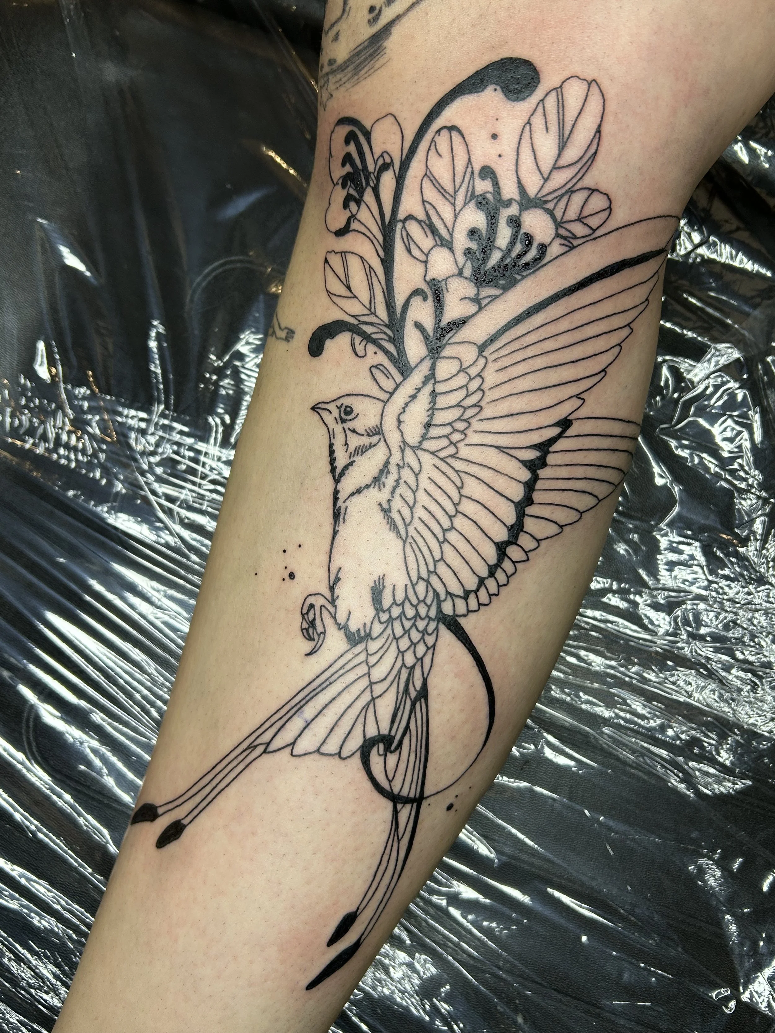 Outline tattoo of a bird perched on a flowering branch with leaves on the upper arm.