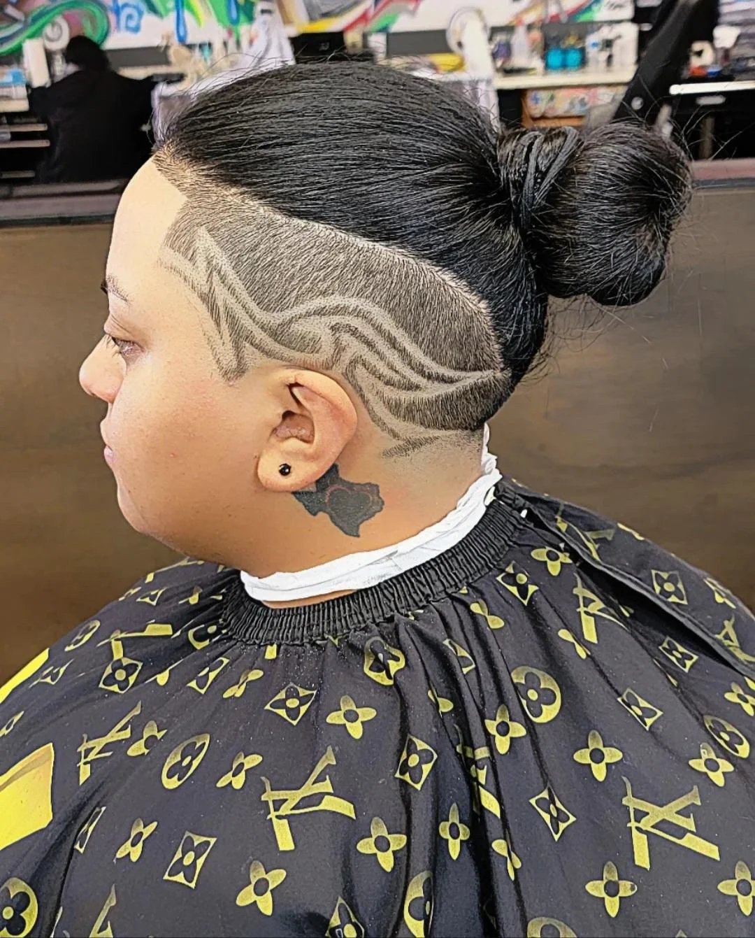 A woman with a hairstyle featuring a shaved design with wavy lines, and her hair styled in a bun. She has a tattoo of the state of Texas on her neck and is wearing a black cape with a gold Louis Vuitton pattern.
