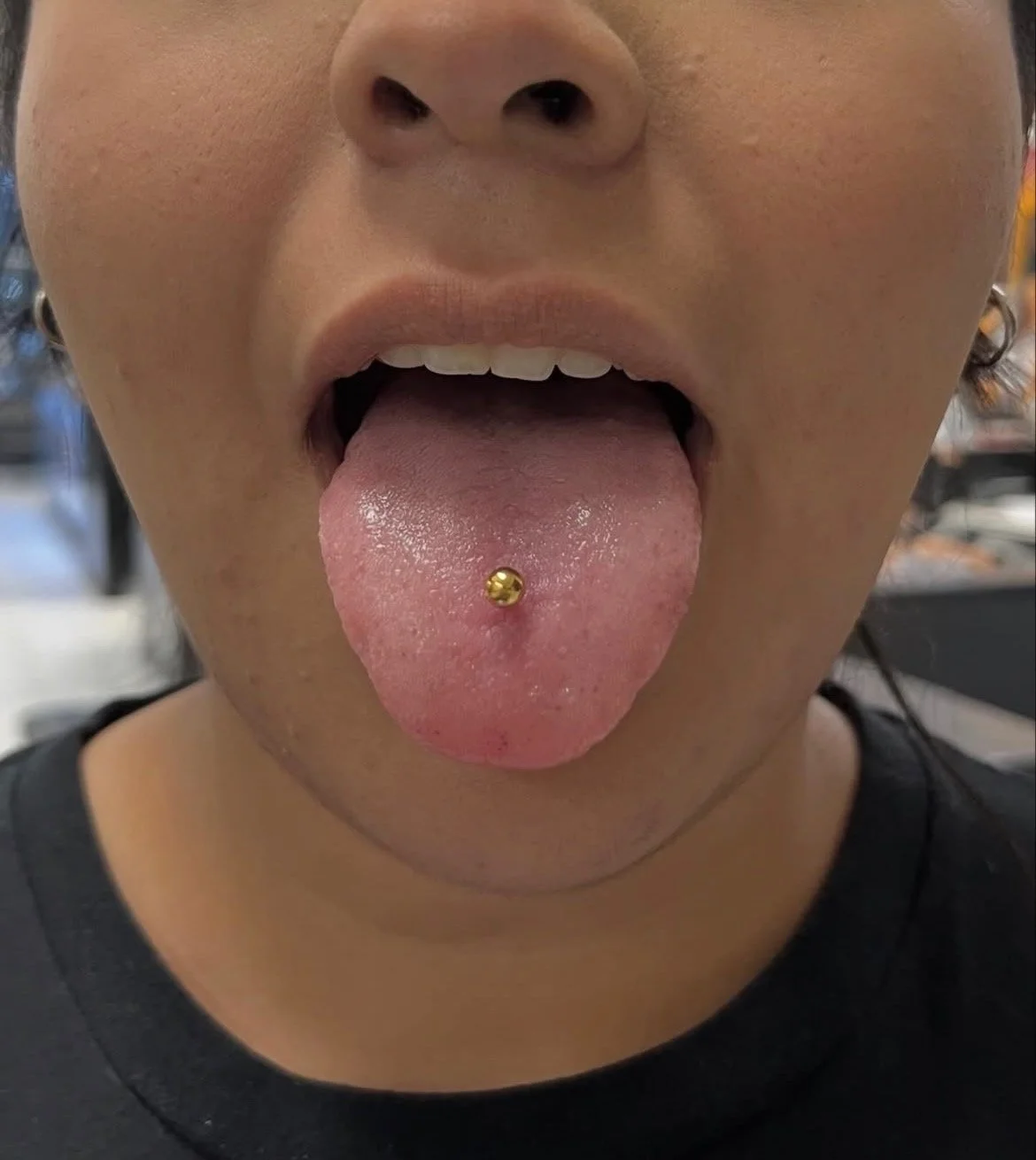 Person sticking out their tongue with a gold tongue piercing in a public setting.