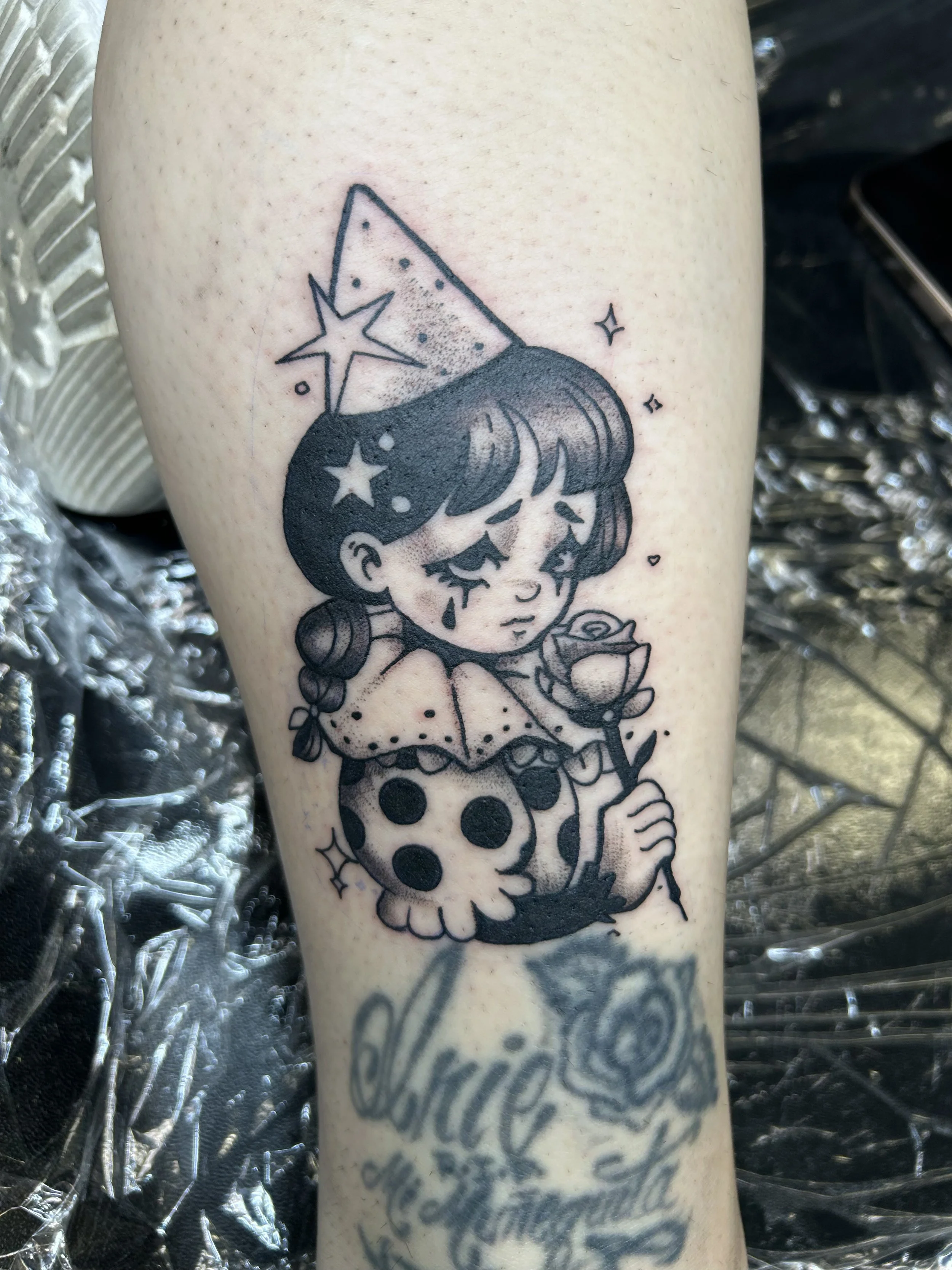 Tattoo of a cartoon girl with short hair, holding a flower, a skull decorated with dots, a star, and other celestial symbols surrounding her.