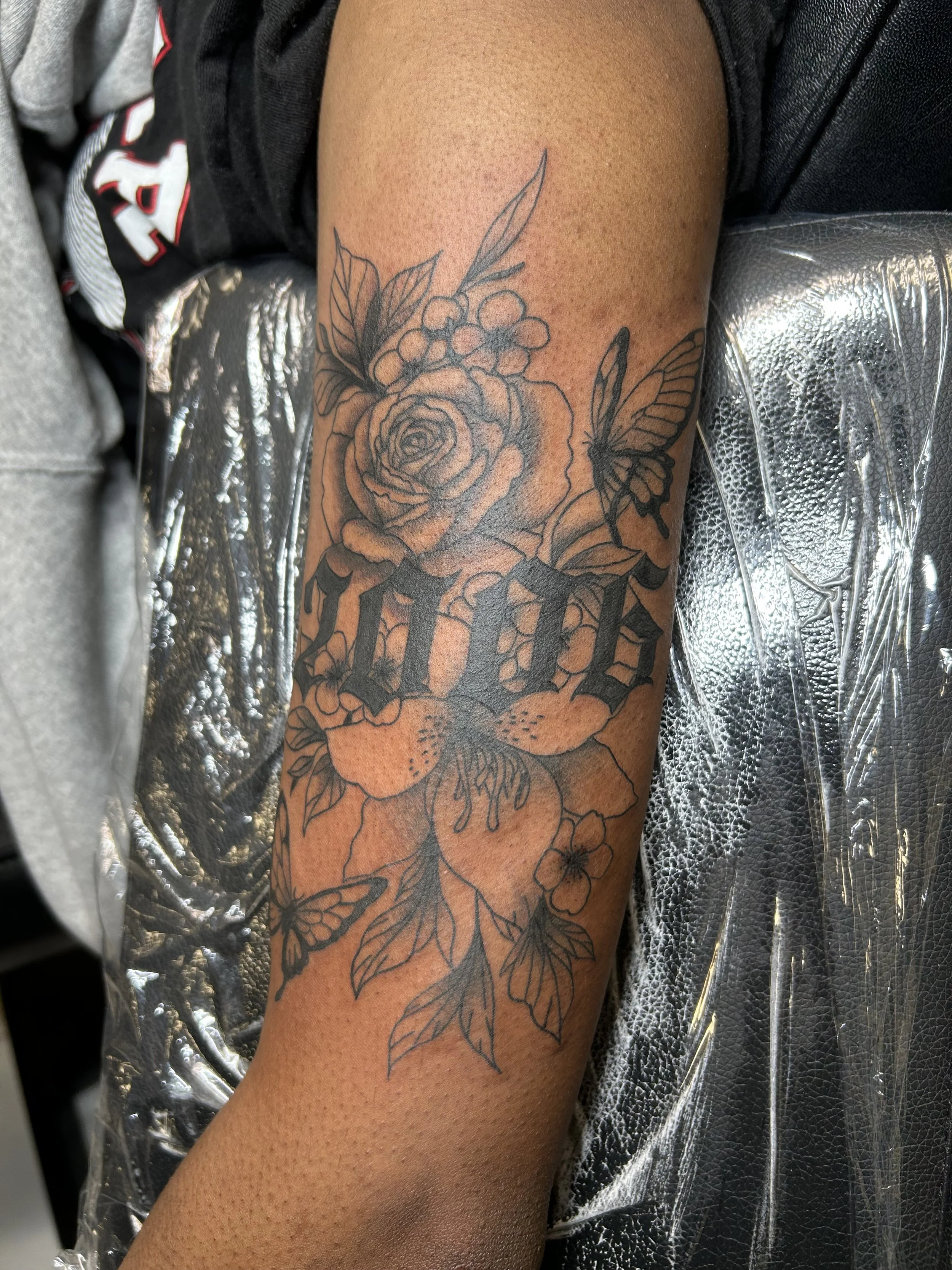 A tattoo on a person's arm featuring a large rose, butterflies, and the word "LOVE" in gothic font, surrounded by leaves and other floral elements.