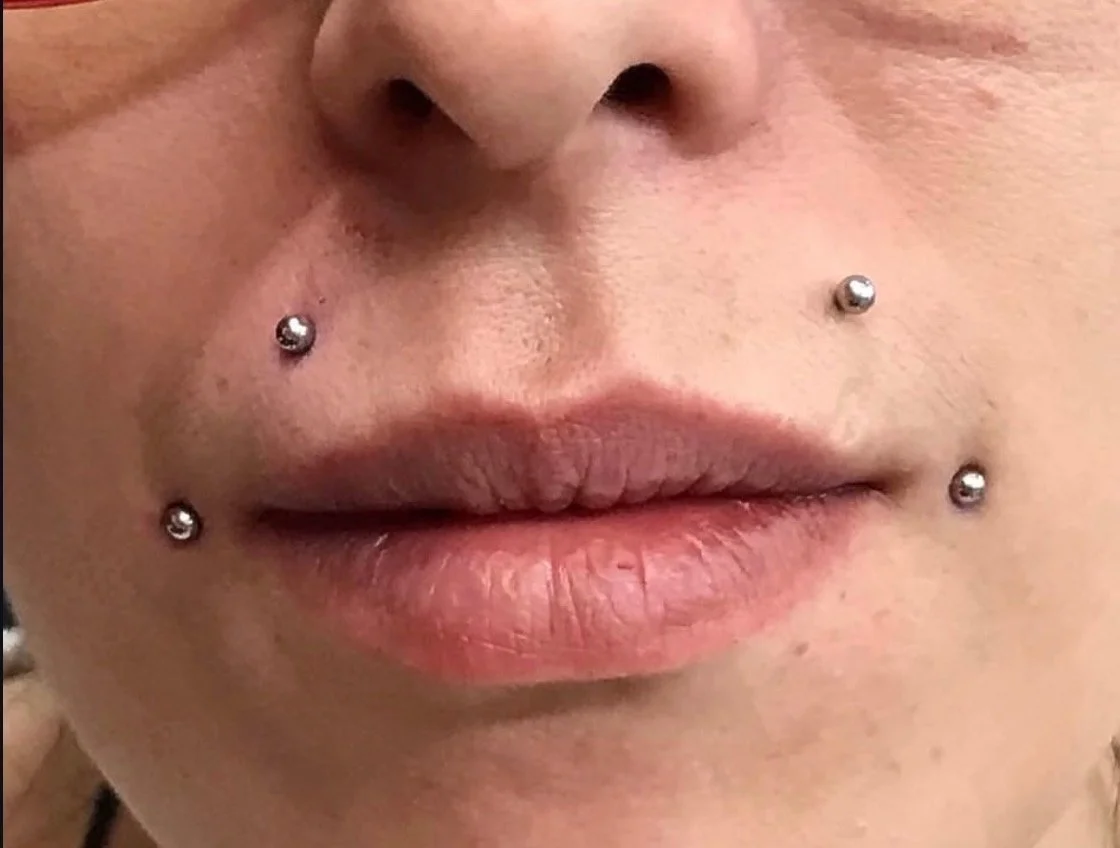 Close-up of a person's lips and face with four silver facial piercings.