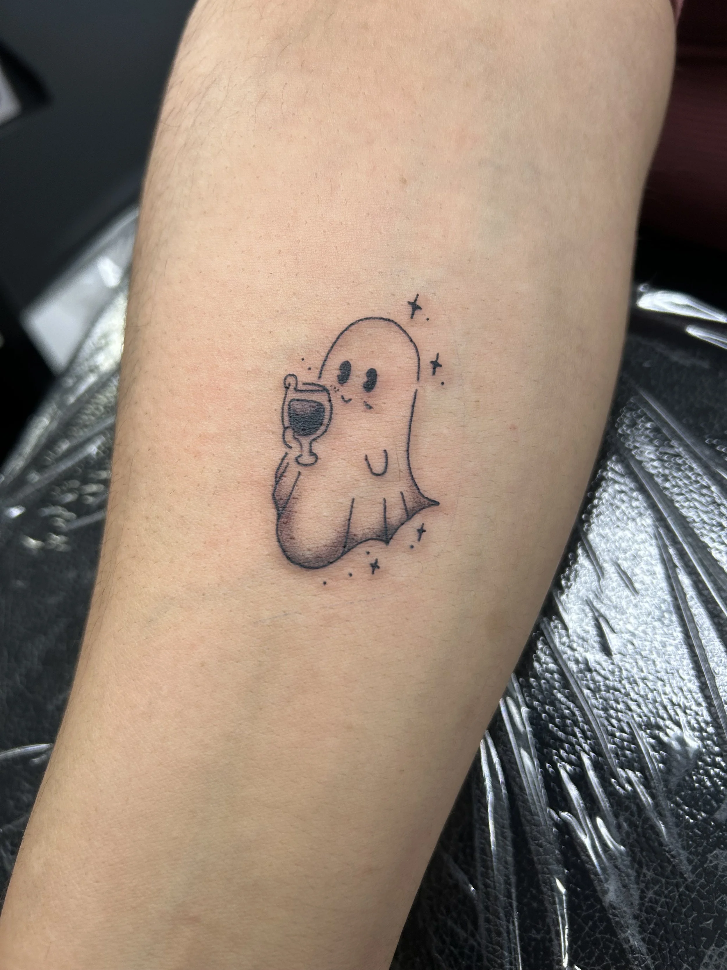 Tattoo of a cute cartoon ghost holding a wine glass, with stars and sparkles around it.