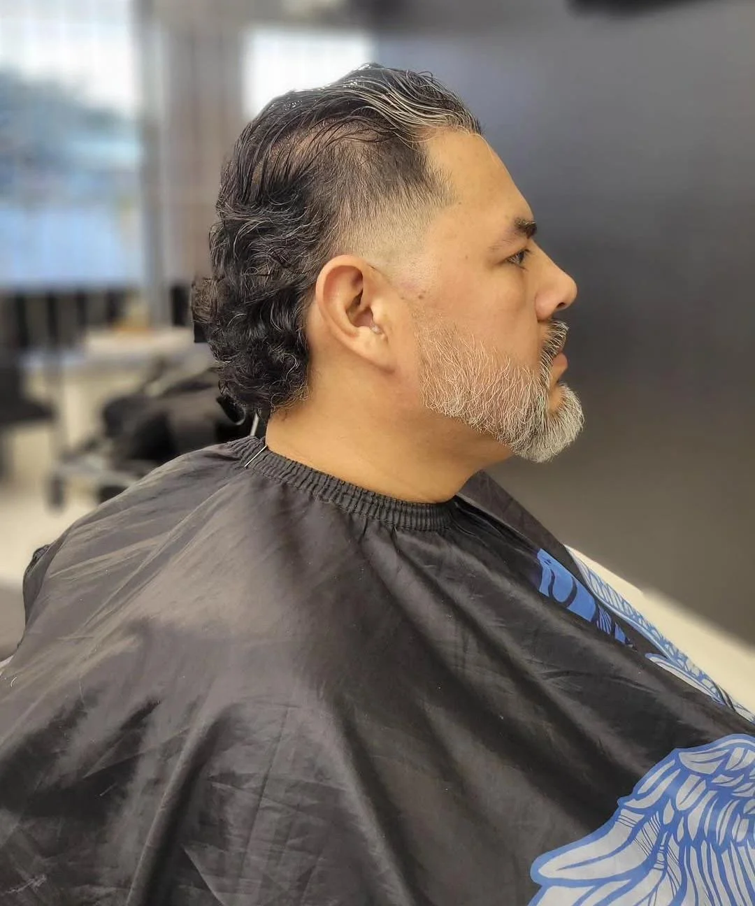 Side view of a man with black and gray slicked-back hair and a full gray beard, sitting in a hair salon chair with a black cape over his shoulders during a haircut.