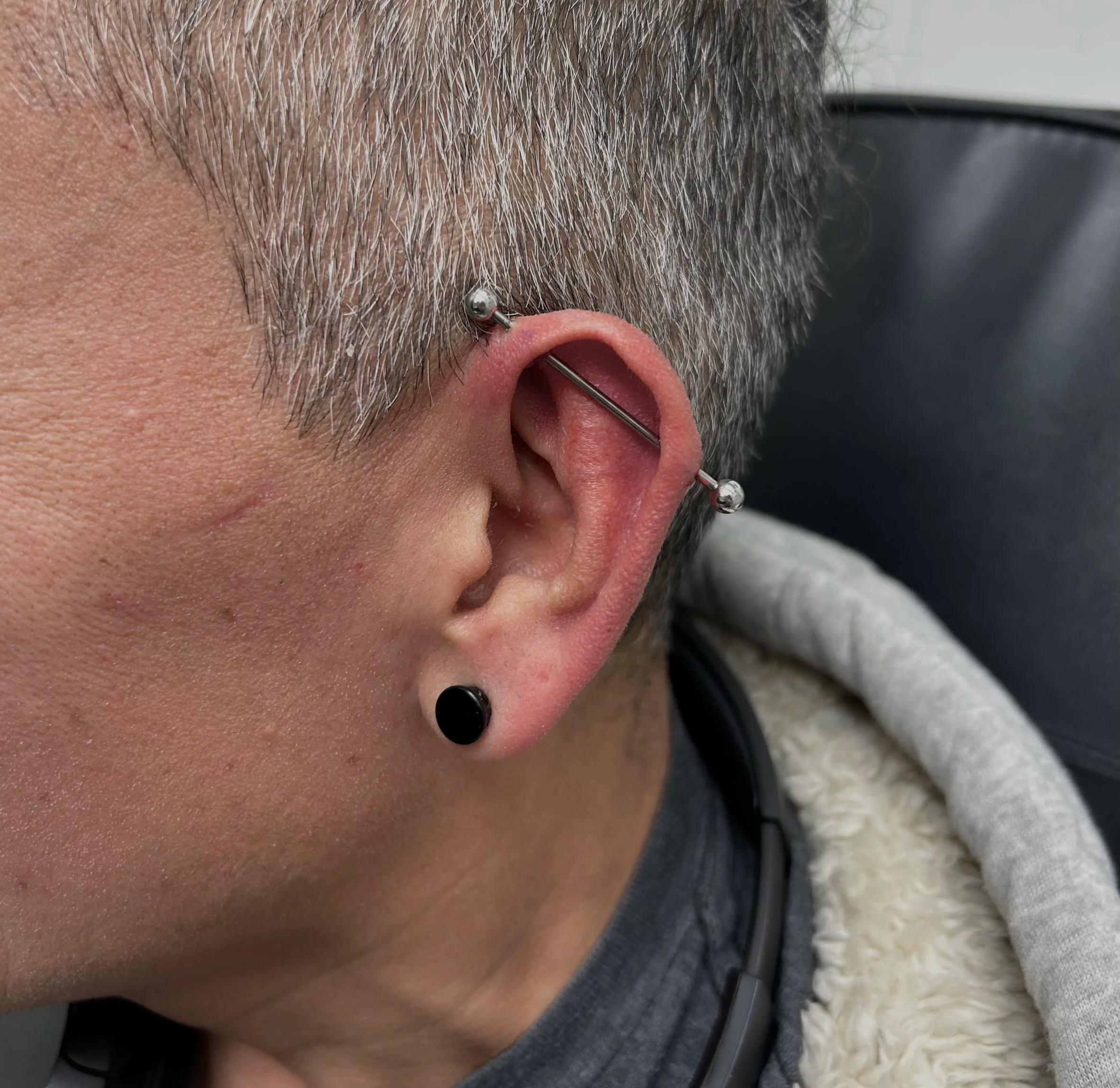 Close-up of a person's ear with multiple piercings, including a curved barbell with metal balls and a black stud earring, gray hair, and skin with some freckles and a scar.