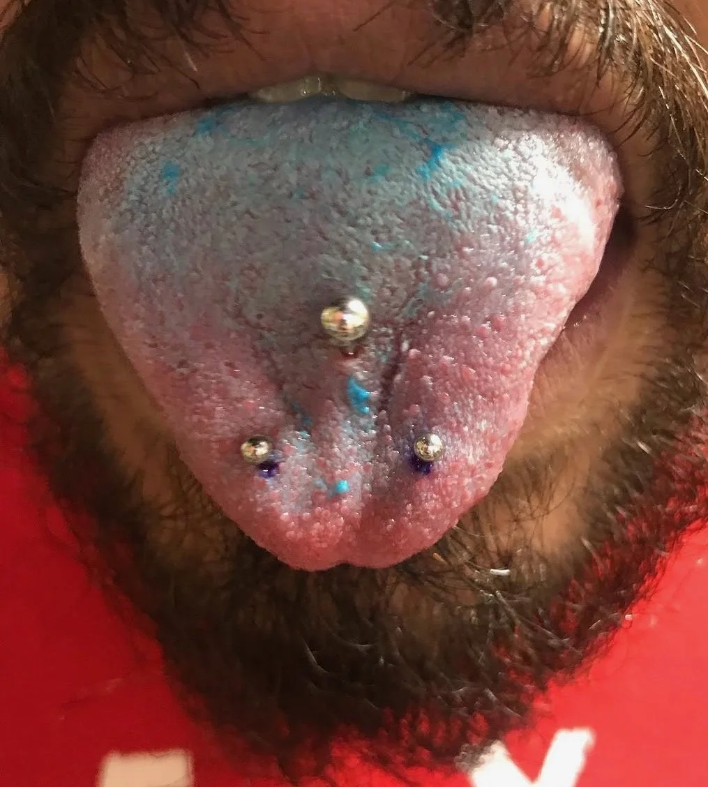 Close-up of a person's tongue with tongue piercings, showing metallic bars and colorful splashes of paint or ink on the tongue surface.