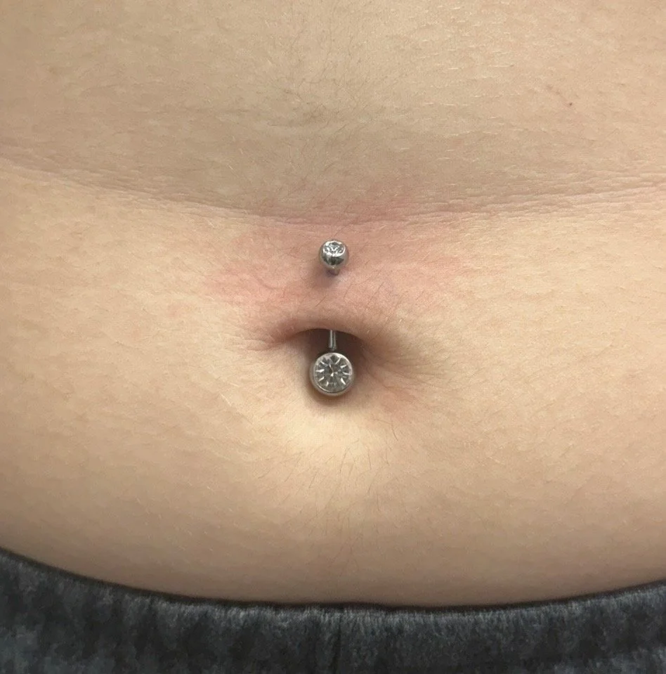 A close-up of a person's navel with a piercing, featuring a small silver ball on top and a larger, circular gem at the bottom.