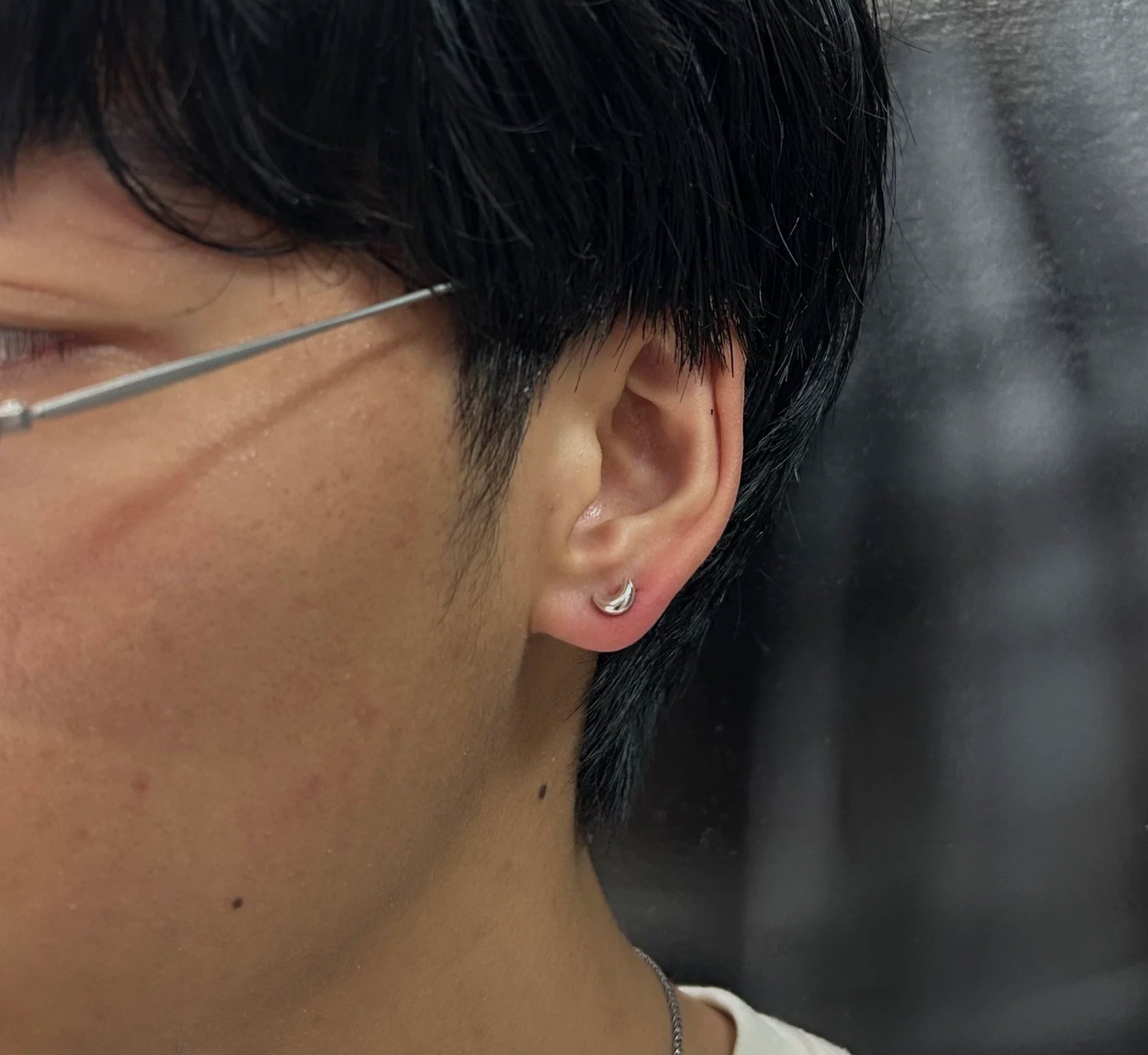 Close-up of a person's ear with a small silver hoop earring, short black hair, and glasses.