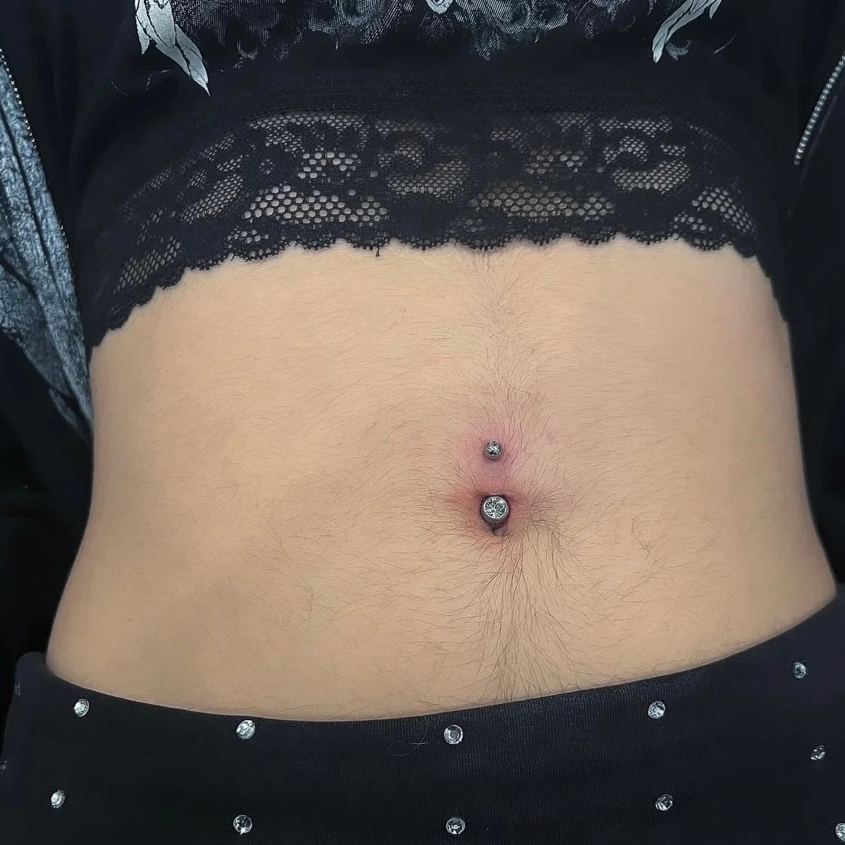 Close-up of a person's bare abdomen with a navel piercing featuring a silver ball and a large gemstone, wearing a black lace top and black pants with small rhinestones.
