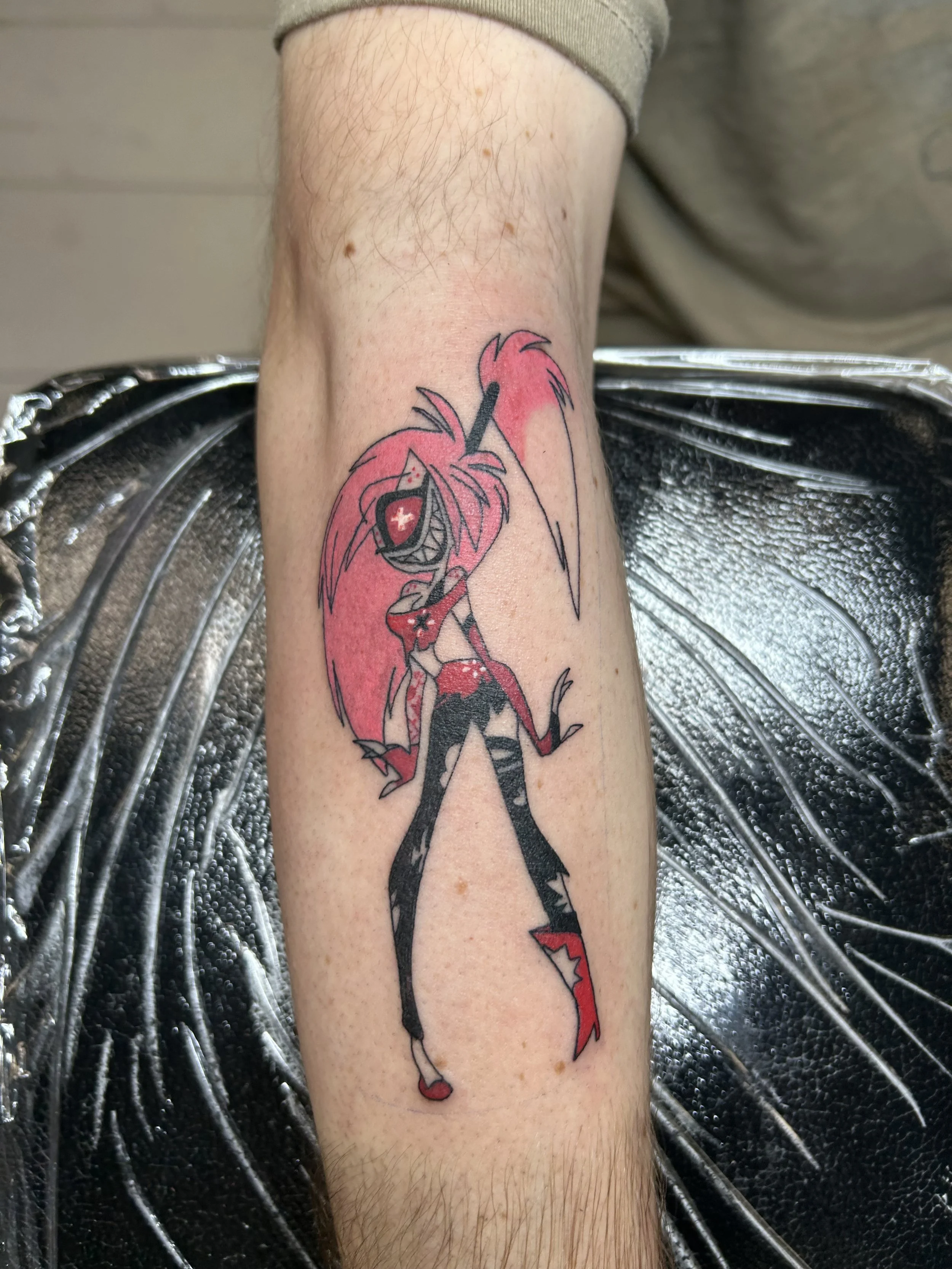 Tattoo of a cartoon girl with pink hair, a mischievous grin, a black and red outfit, and a long red and pink tail, on a person's forearm.