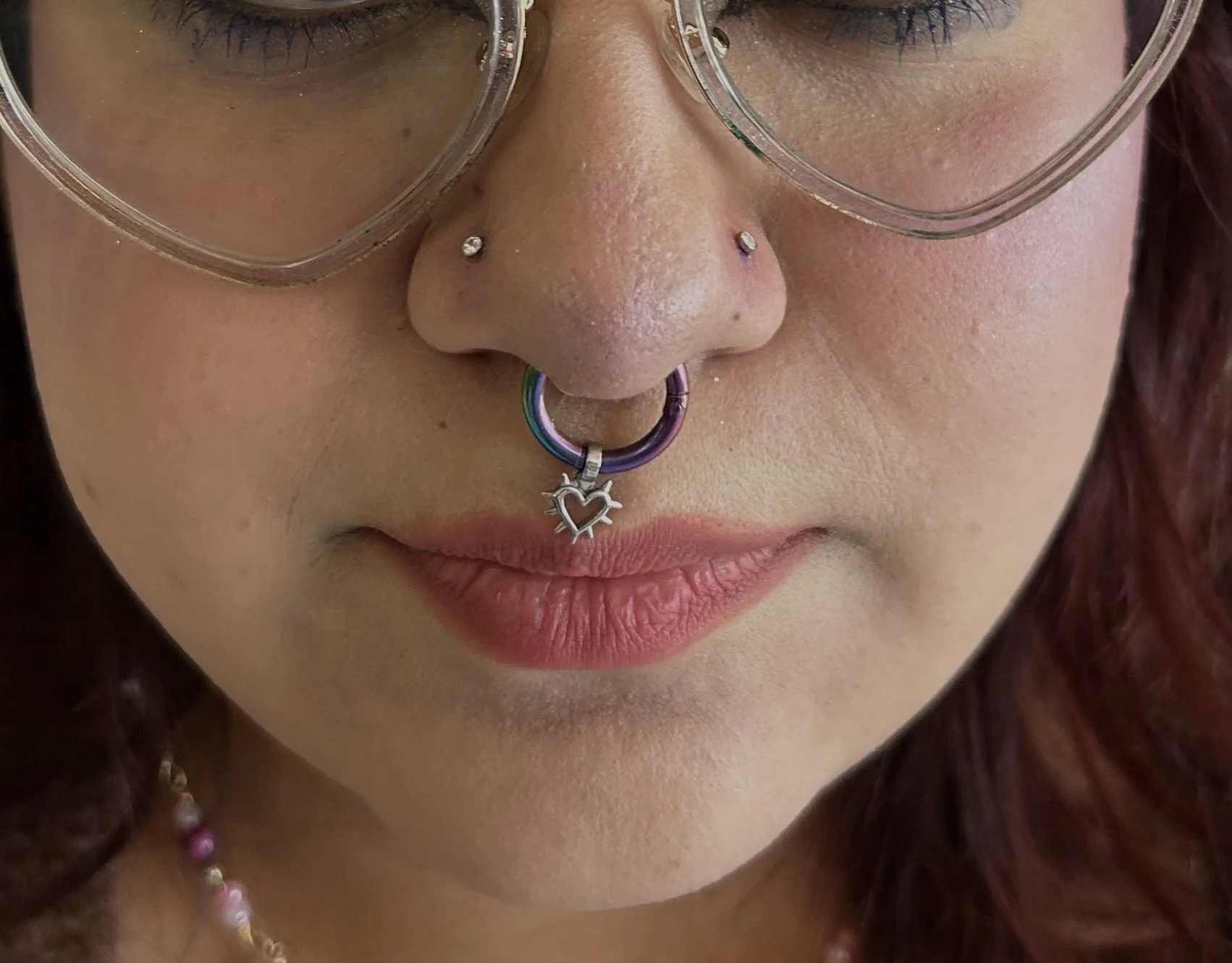 Close-up of a person with glasses, nose piercings including a septum ring with a heart-shaped charm, and wearing a pearl necklace. The person has medium skin tone and reddish-brown hair.