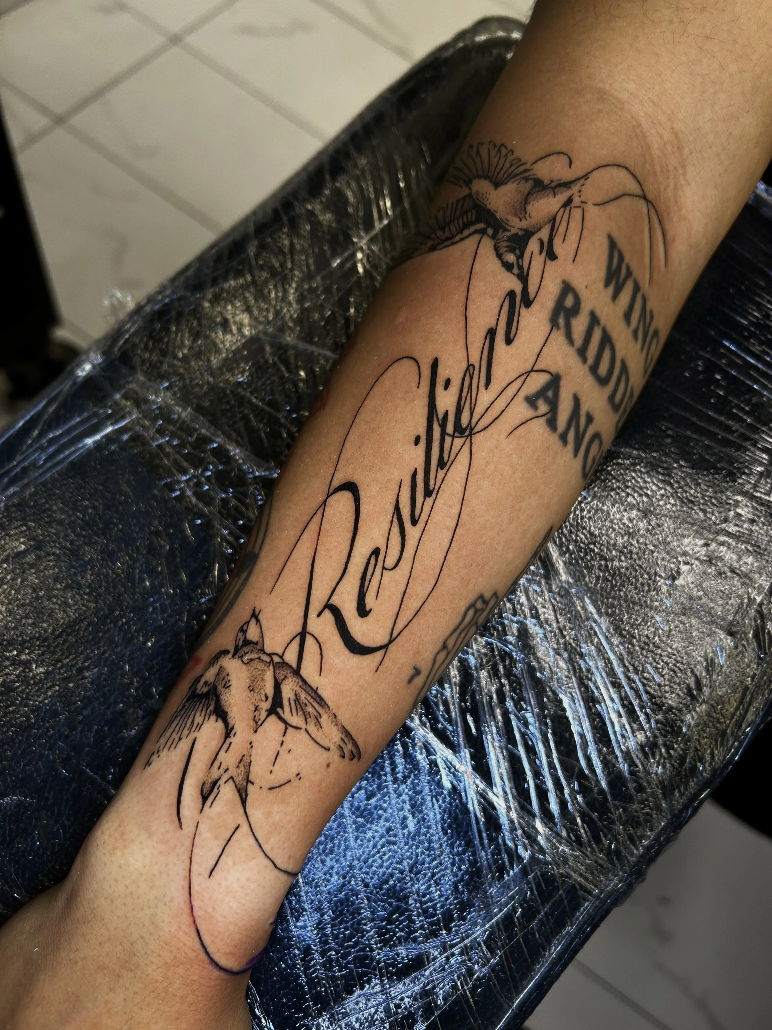 A person's forearm with a new tattoo of a large flower and cursive script, wrapped in plastic wrap and resting on a textured, shiny surface.