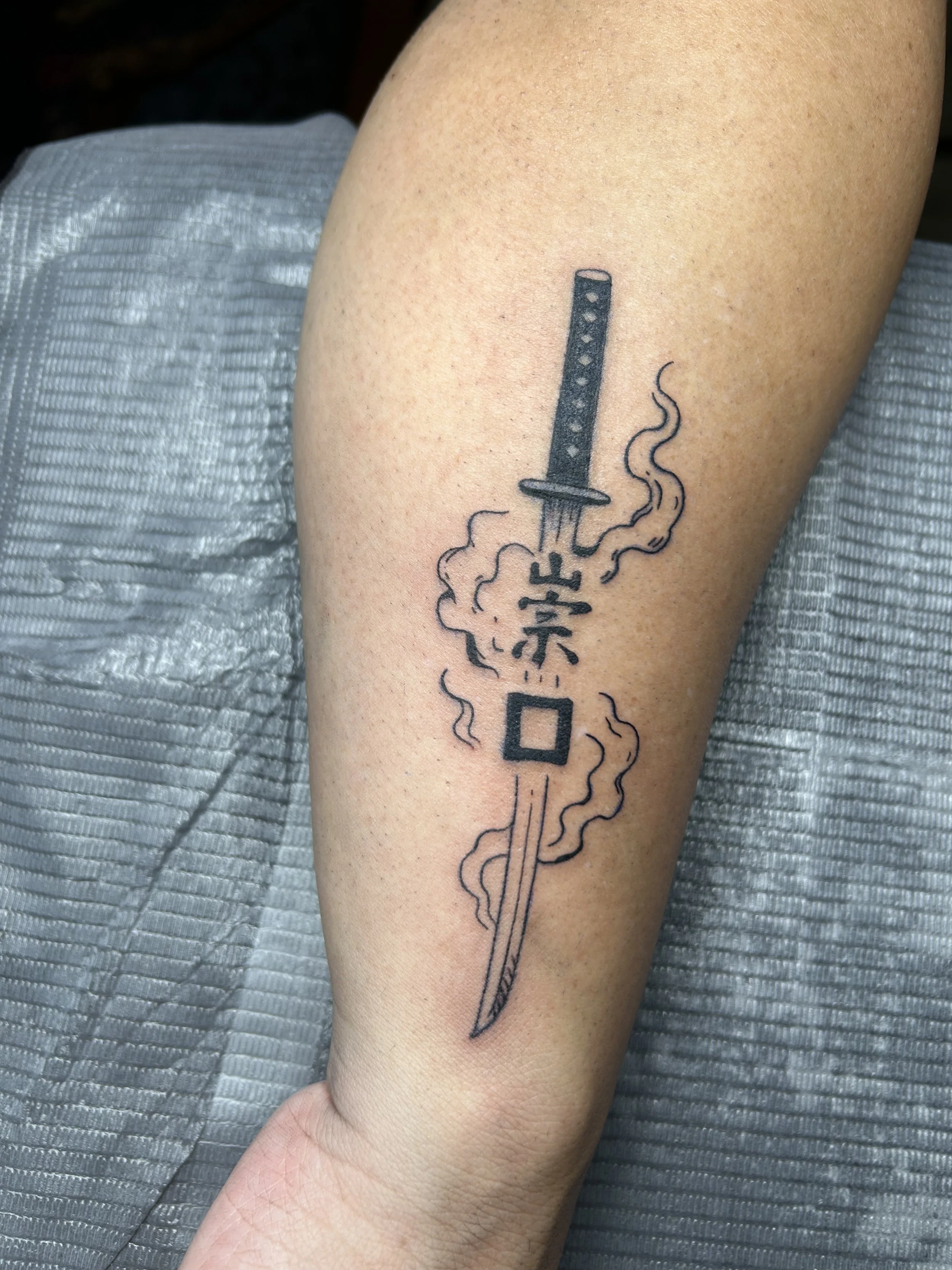 Tattoo of a sword with smoke and Japanese characters on a person's arm.