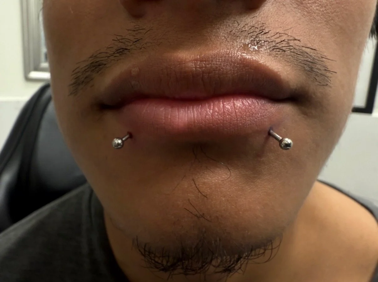 Close-up of a person's lips with two lip piercings and facial hair.