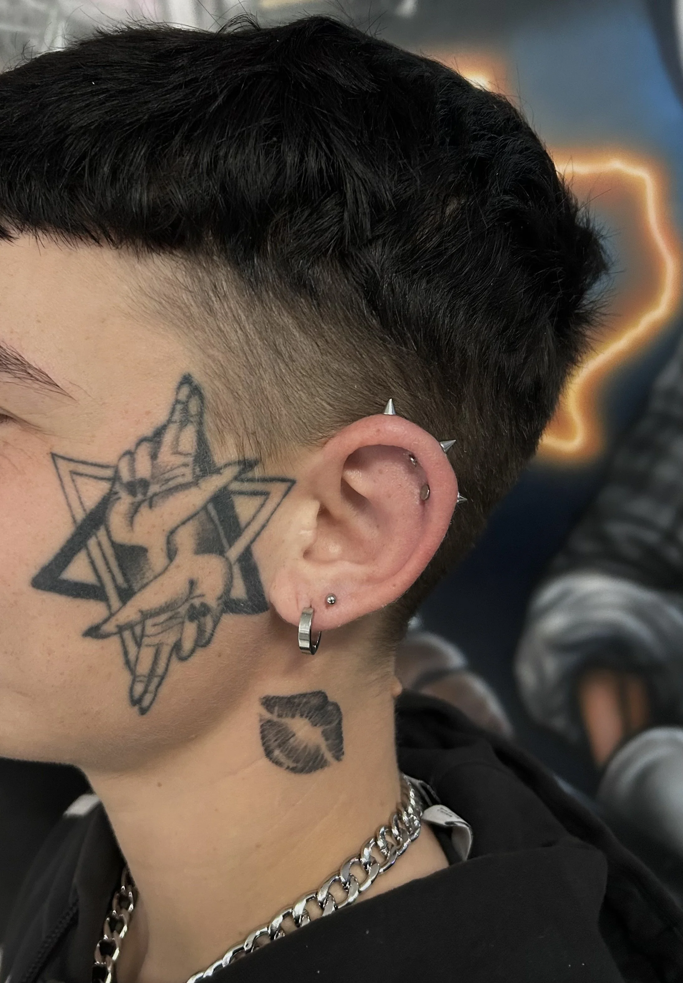 A person with short, dark hair and multiple tattoos near the ear, including a hand holding a pencil and a pair of lips, wearing multiple earrings and a silver chain necklace.