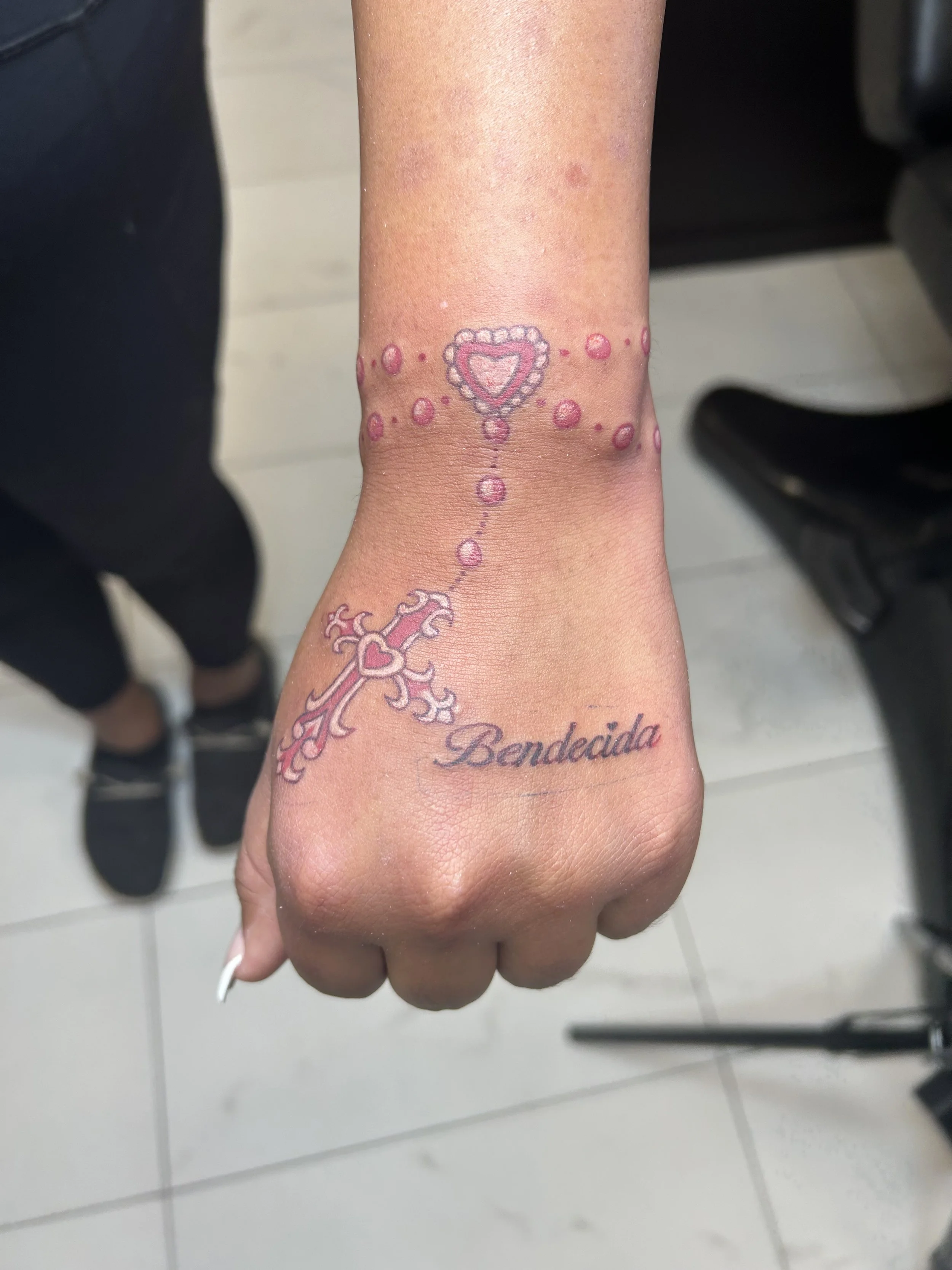 Tattoo on a person's hand and wrist featuring a decorative cross, pink and purple beads, a heart, and the word 'Bendecida' in cursive.