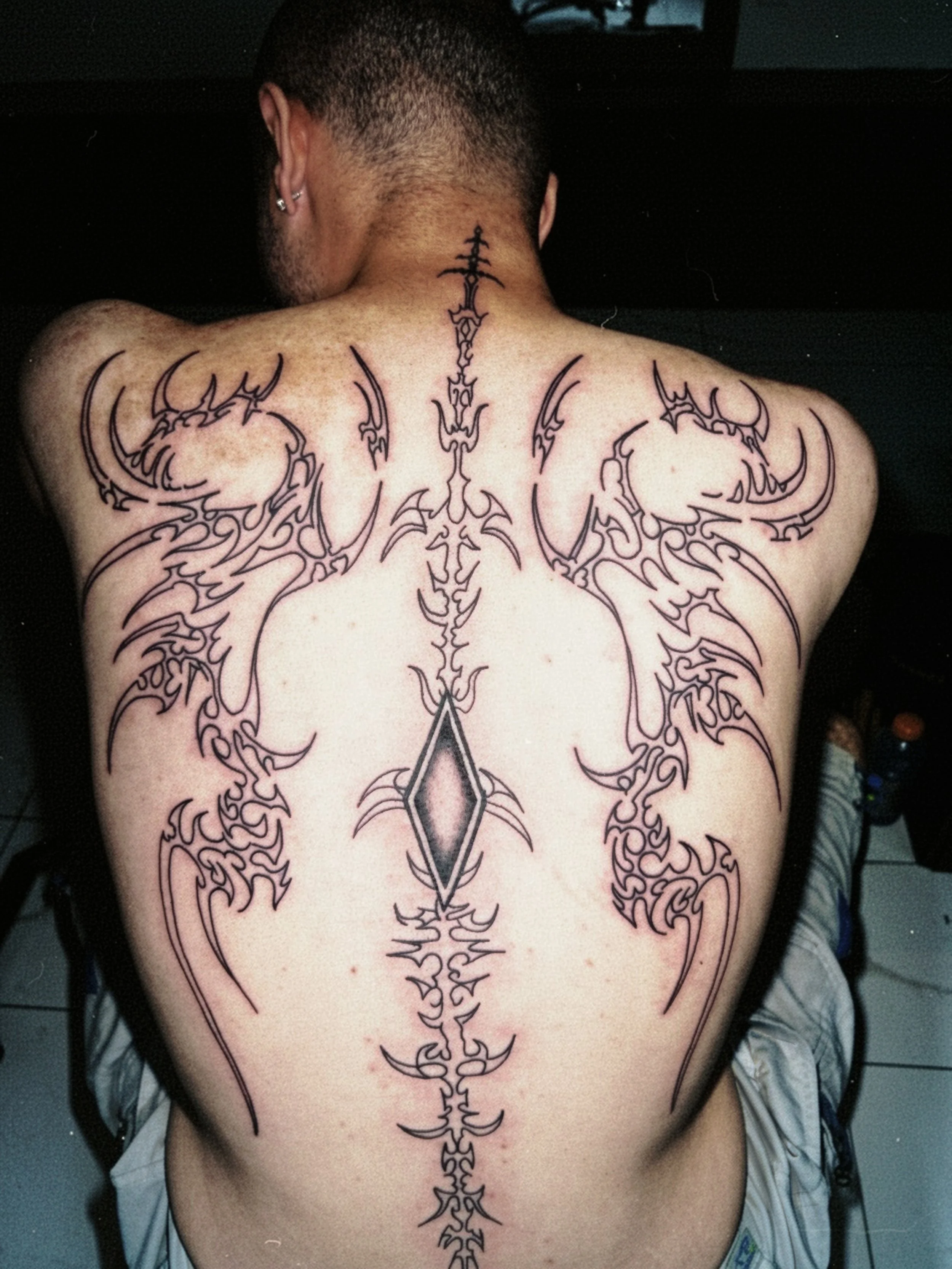 A person with short dark hair has an intricate black tattoo that runs down the center of their back, from the neck to the lower back. The tattoo features symmetrical, ornate, tribal-style patterns with sharp, pointed edges.