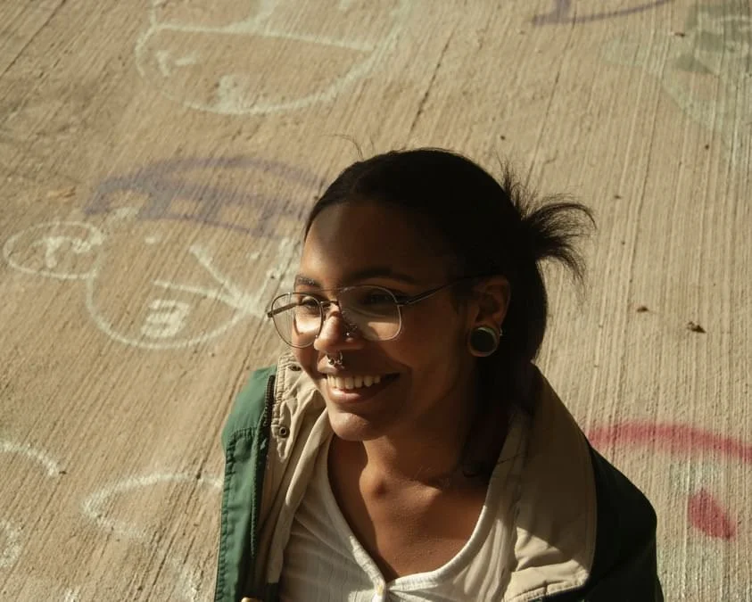 A woman with glasses and dark hair smiling outdoors on a sidewalk with chalk drawings.