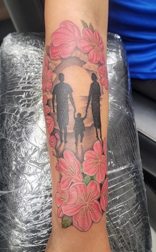 Tattoo of a silhouette of a family holding hands, walking on a beach with pink flowers surrounding the scene.
