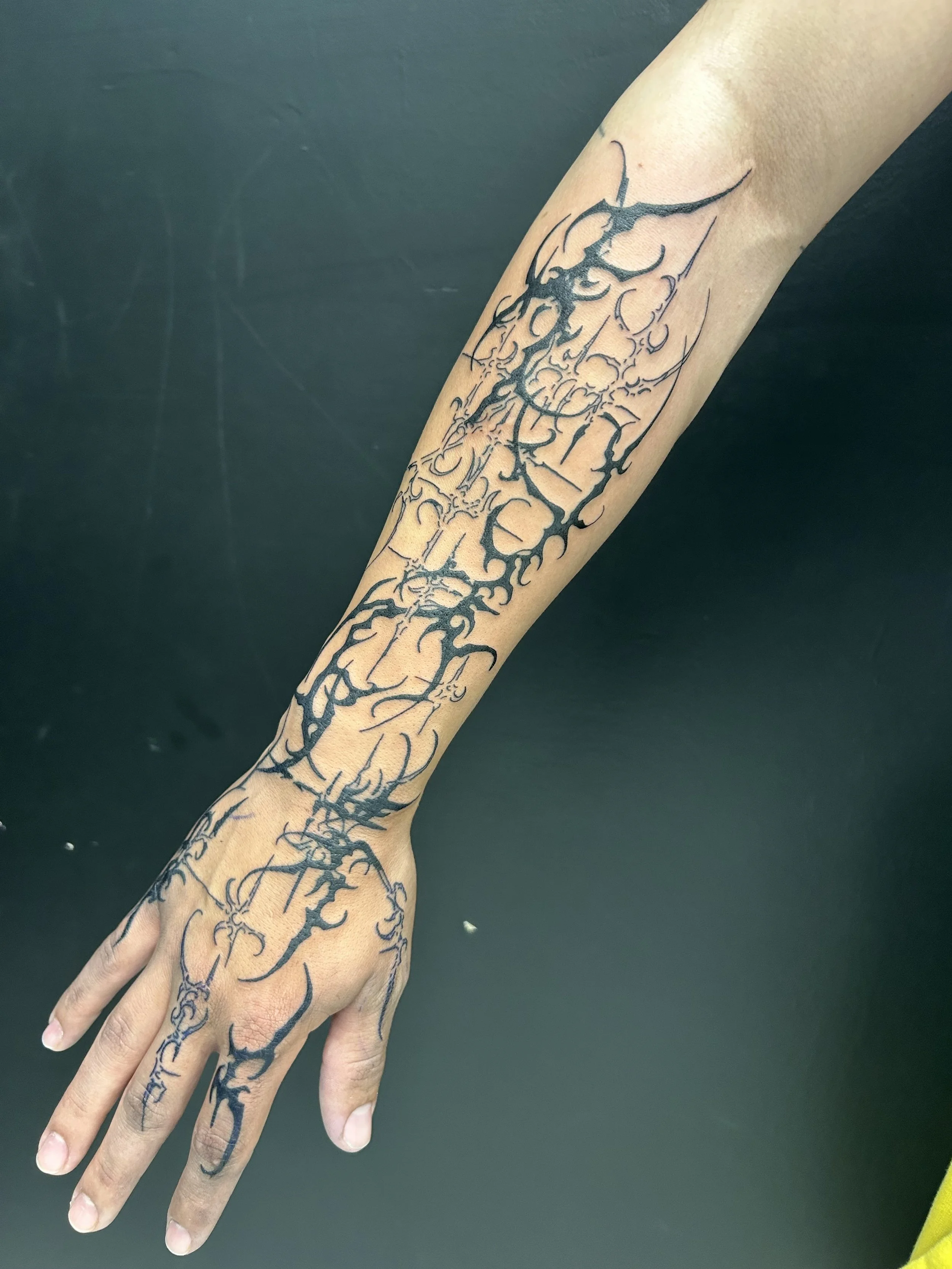 Tattoo of a dark, abstract design extending from the wrist to the upper forearm, inspired by tribal or gothic art styles, with intricate line work and sharp, flowing patterns.