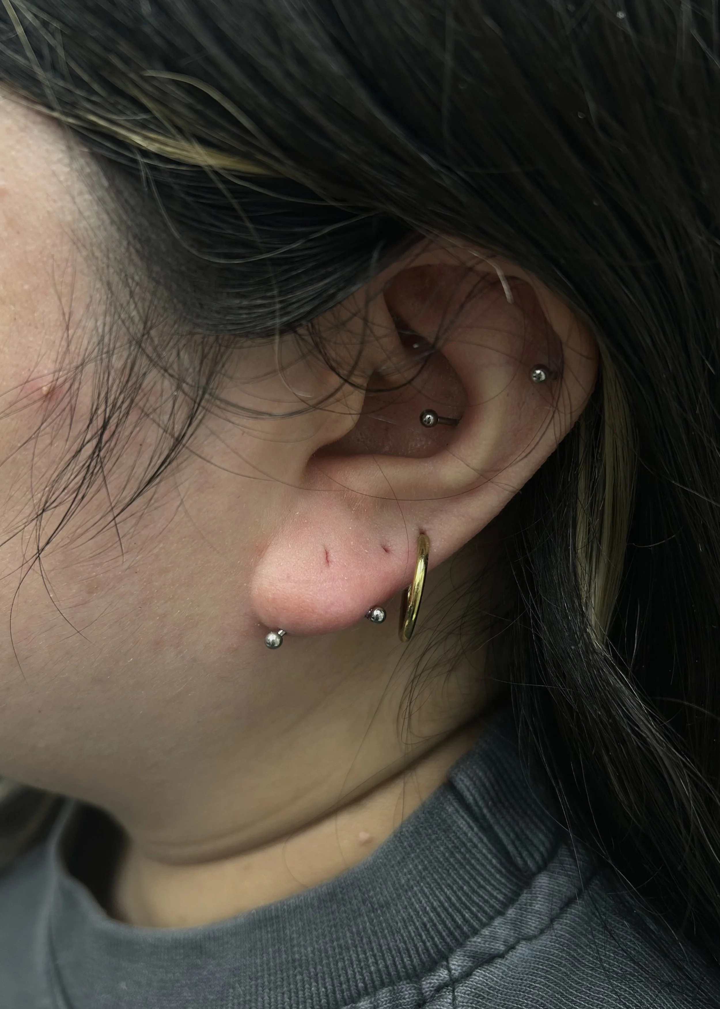 Close-up of person's ear with multiple piercings, including a cartilage hoop, a hoop in the earlobe, and a barbell in the helix, with dark hair partially covering the ear.