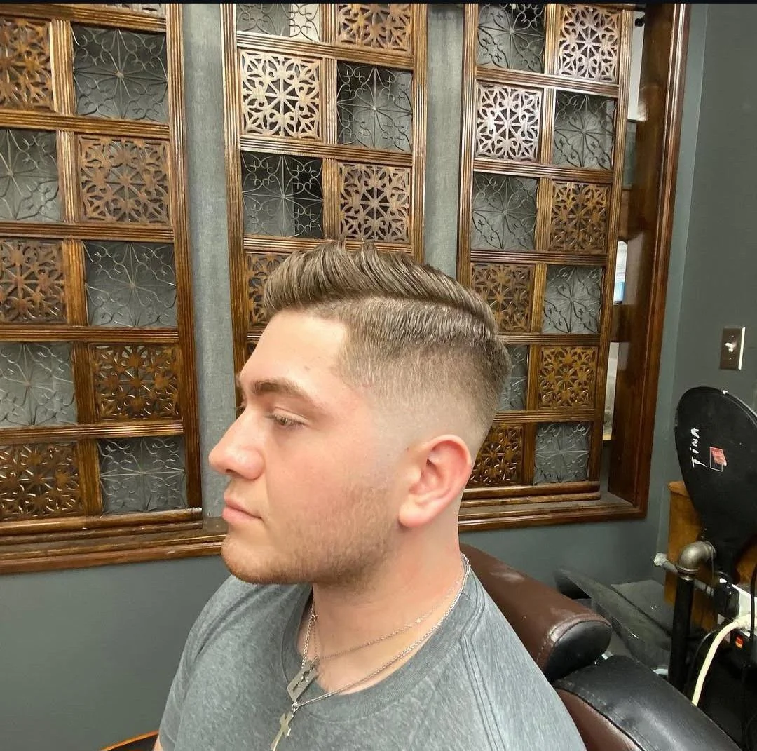 A young man with a fresh, slicked-back hairstyle and a fade haircut sitting in a barber shop chair, with decorative wooden panels in the background.