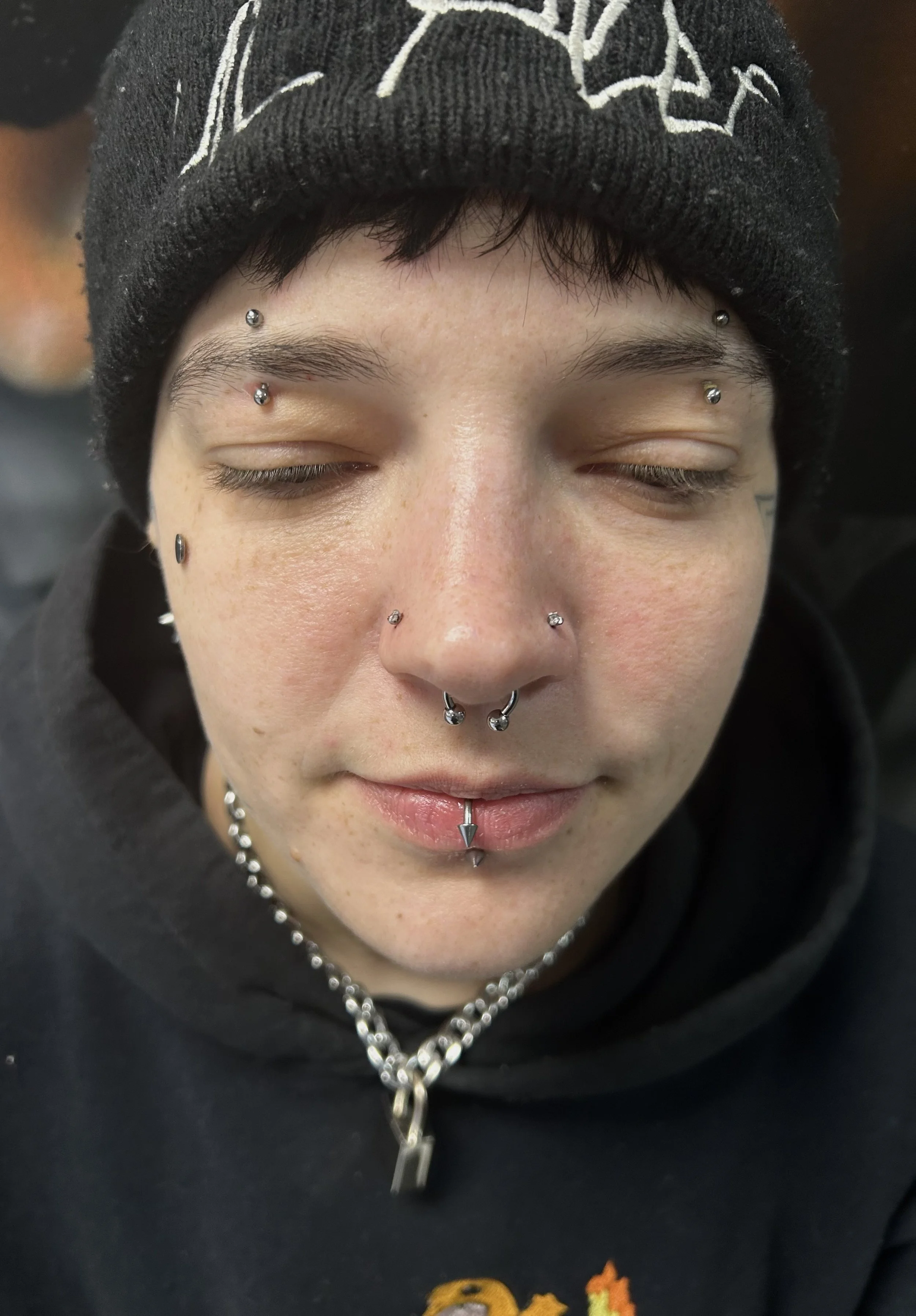 Close-up of a person's face with multiple facial piercings, including septum, nostril, eyebrow, and lip piercings. The person is wearing a black beanie hat and a black hoodie with cartoon graphics, along with a chain necklace.
