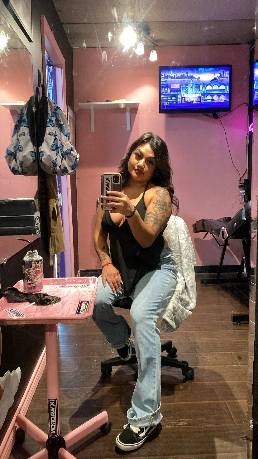 A woman taking a mirror selfie in a room with pink walls, wearing a black tank top, light blue jeans, and black sneakers. There are various objects including a pink table, a black chair, hanging bags, a TV screen on the wall, and a window with purple lighting.